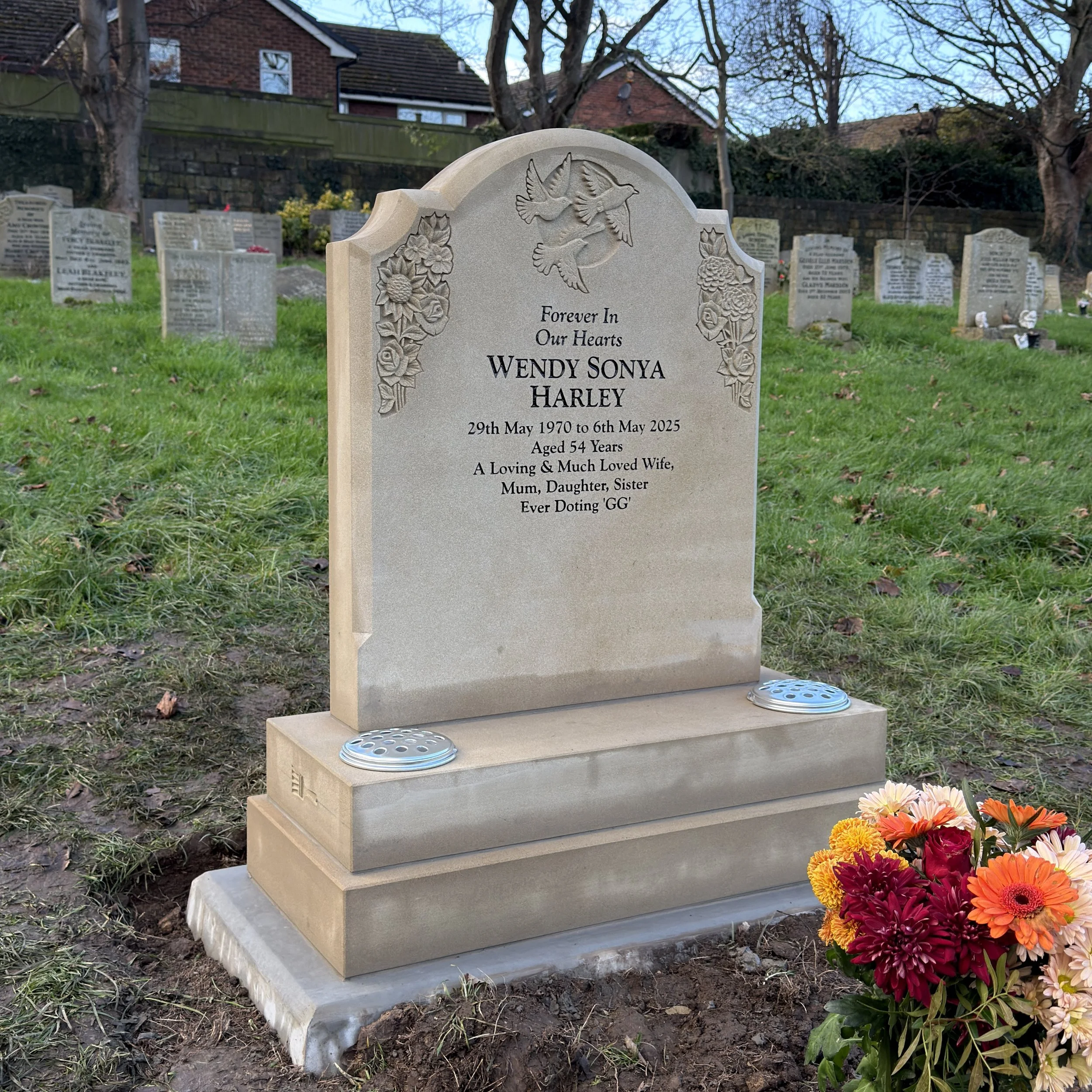 A gravestone in a cemetery with engraved floral decorations and a flying bird at the top. It bears the name Wendy Sonya Harley, who lived from May 29, 1970, to May 6, 2025, and describes her as a loving wife, mum, daughter, and sister. There are fresh flowers at the base of the gravestone.