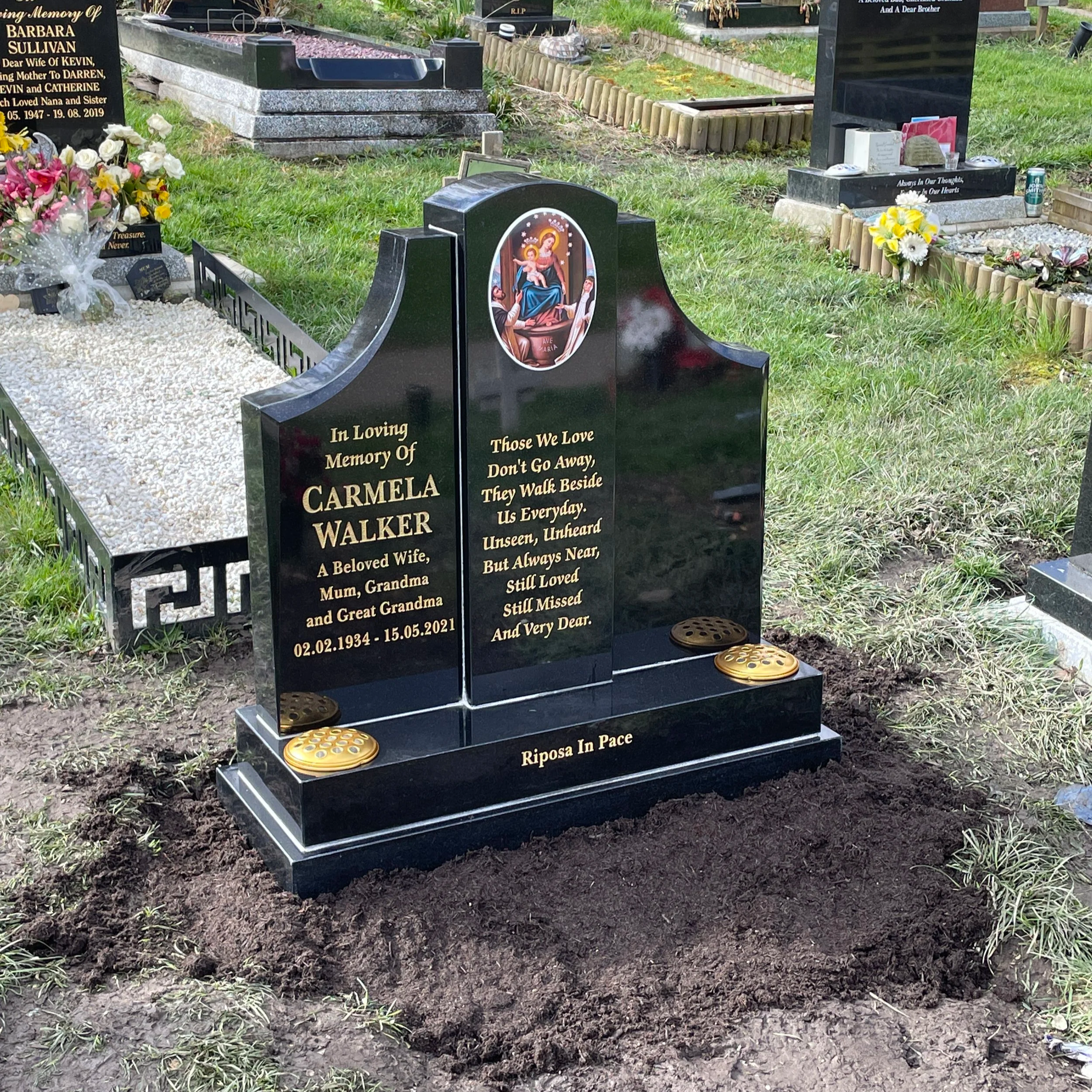 Black granite gravestone with gold lettering for Carmela Walker. The gravestone features an oval picture of the Virgin Mary and baby Jesus at the top. The inscription includes her name, relation, birth and death dates, and a heartfelt message. A small incense holder and a decorative item are placed on the base. The surrounding area has grass, flowers, and other gravestones.