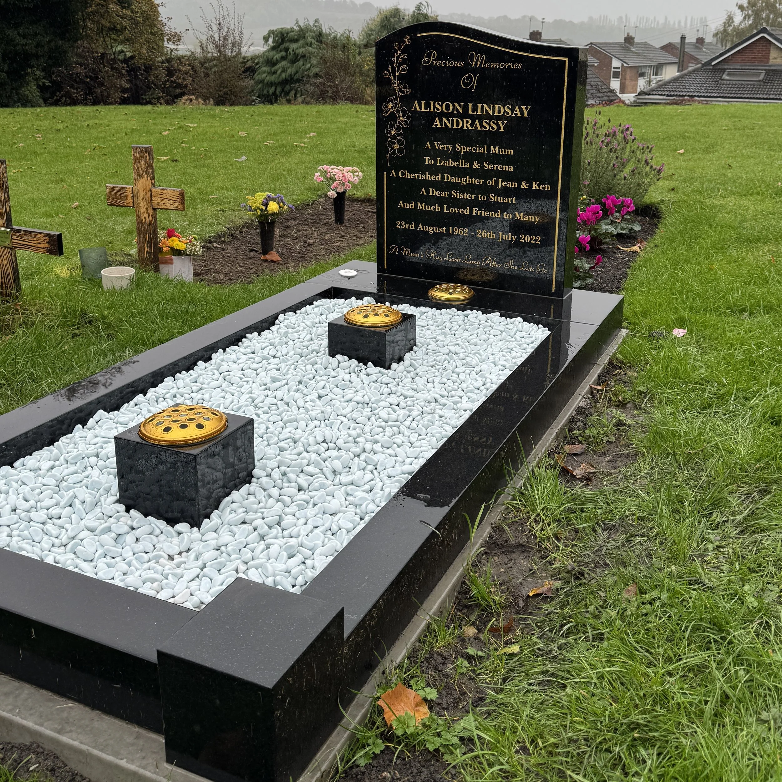 A black granite gravestone with gold detailing and inscription, surrounded by white gravel and two black lanterns. Small floral arrangements and cross markers are in the background on a grassy area with houses and trees.