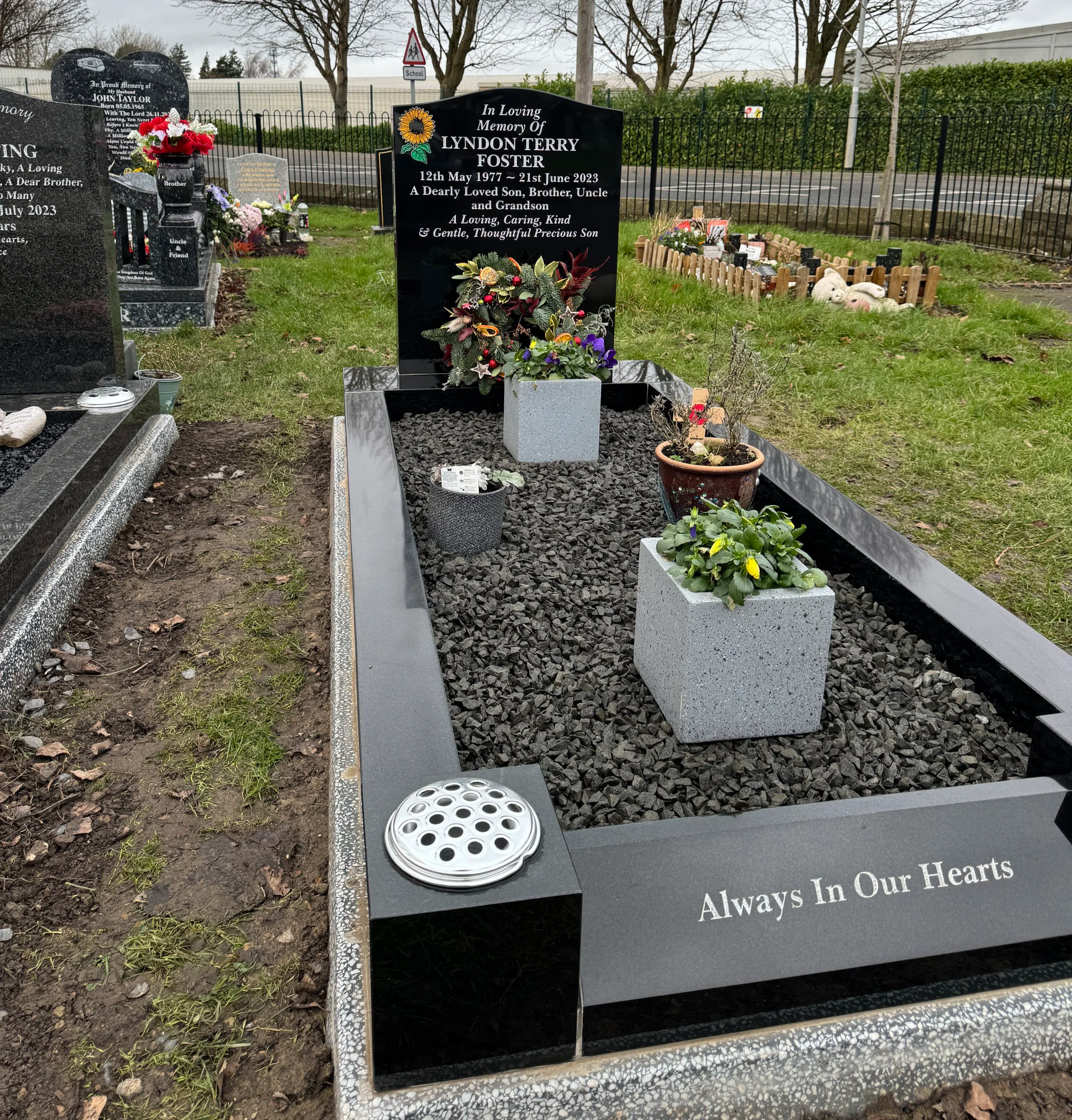 Gravestone in a cemetery with black polished stone, floral arrangements, and potted plants. The headstone reads, "In Loving Memory of Lyndon Terry Foster, 12 May 1977 - 21st June 2023, A Dearly Loved Son, Brother, Uncle and Grandson, A Loving, Caring, Kind & Gentle, Thoughtful Precious Son." The border of the grave is decorated with small stones, and the side of the border has the phrase "Always In Our Hearts" engraved.