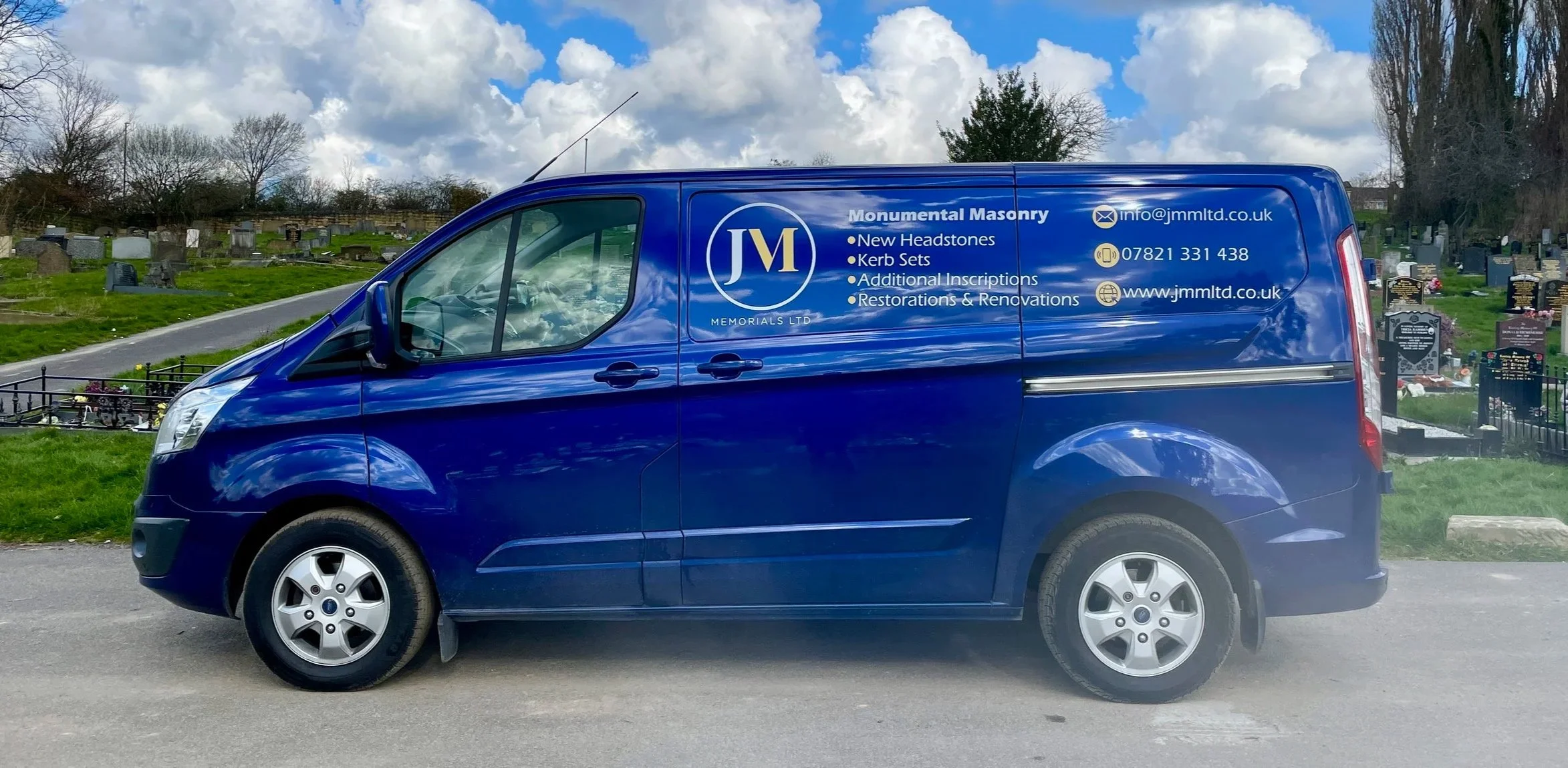 Blue delivery van advertising Monumental Masonry services parked on a street near a cemetery with graves and tombstones in the background.