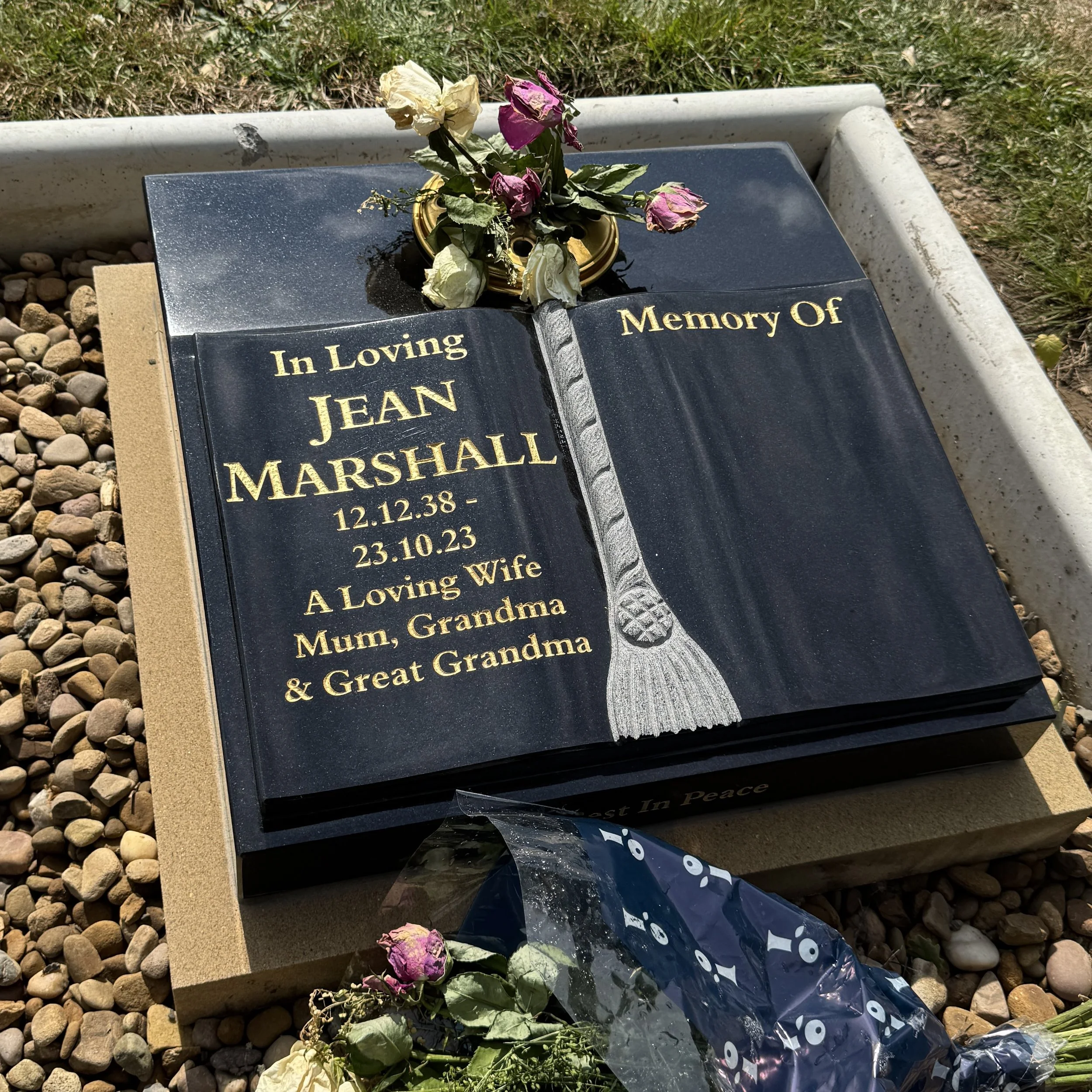 Black granite memorial with gold lettering dedicated to Jean Marshall, includes dates of birth and death, surrounded by flower arrangements and dried roses, set on a concrete base with small gravel ground.