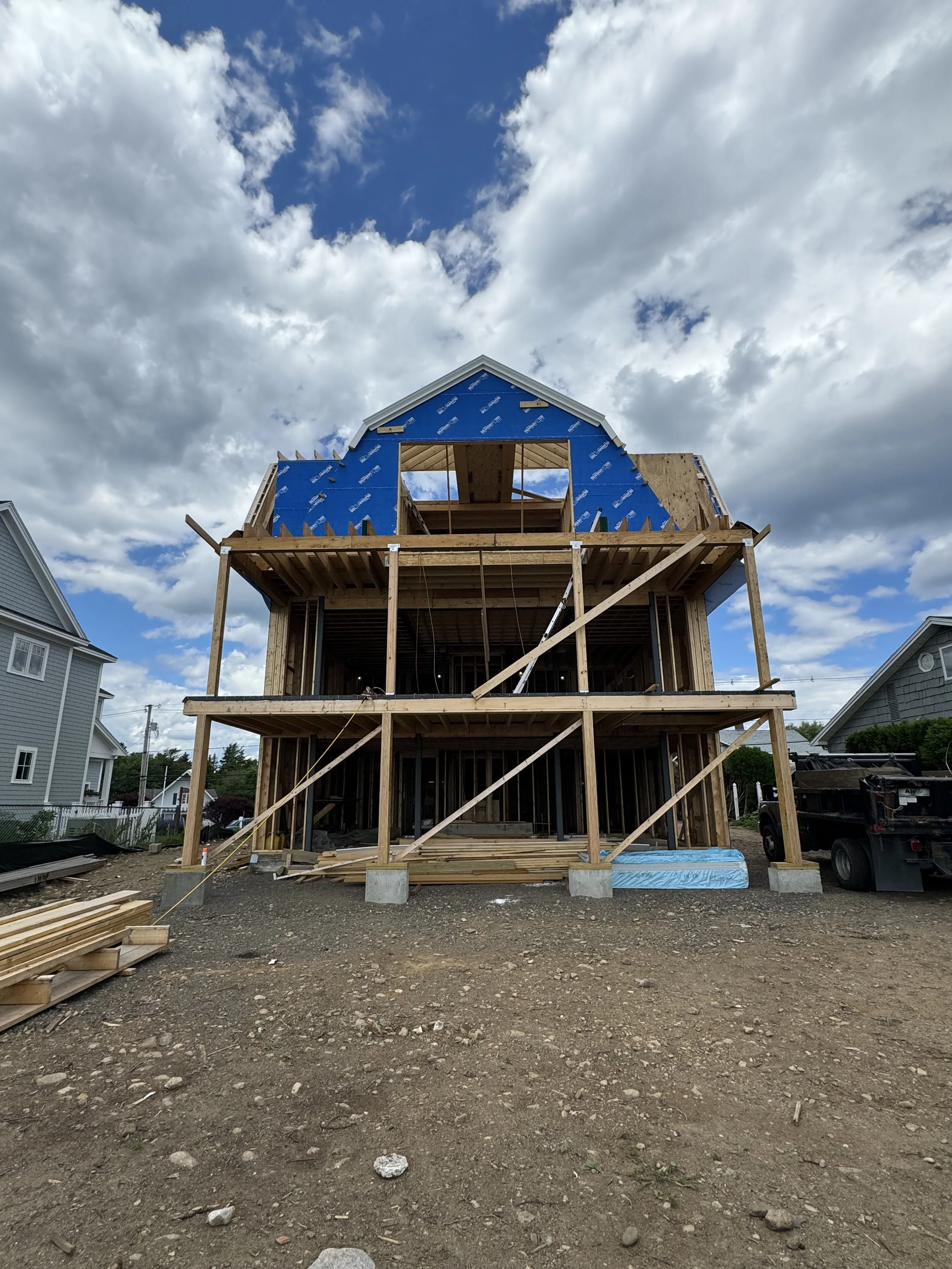 Under-construction wooden house with blue sheathing, three stories tall, with scaffolding, supports, and building materials visible, set against a partly cloudy sky.