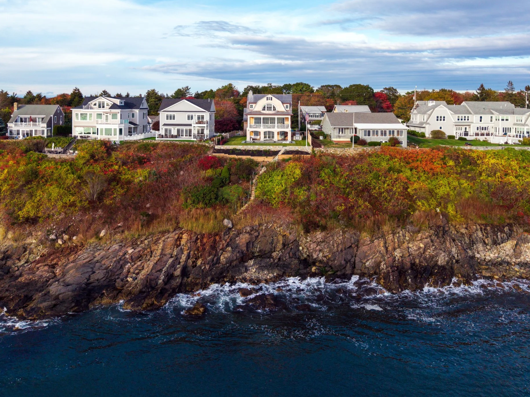 Houses on a cliff overlooking the ocean with rocky shoreline, colorful trees, and cloudy sky.