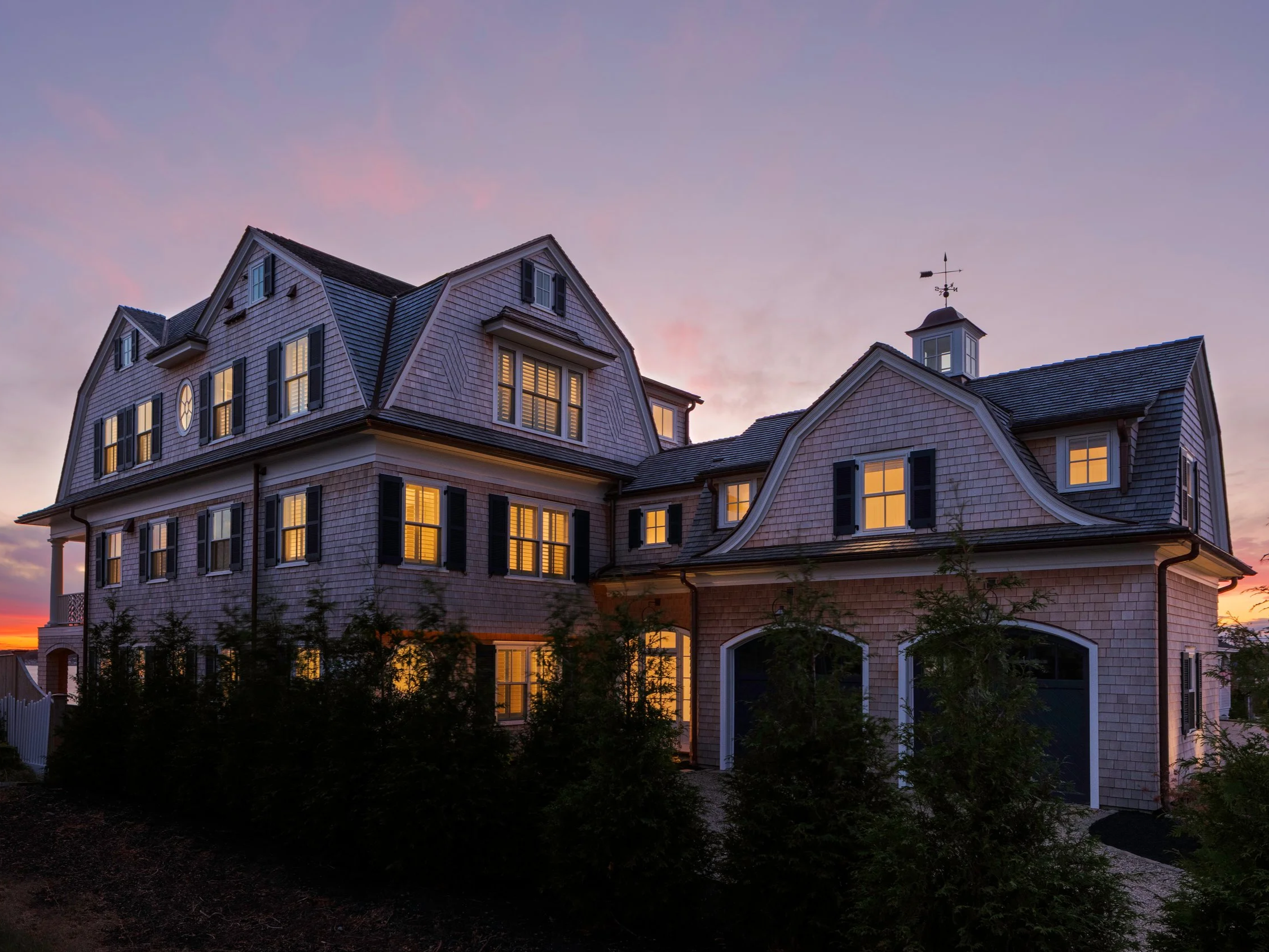 Large house in york maine with multiple floors and windows at sunset, with some lights on inside, surrounded by bushes.