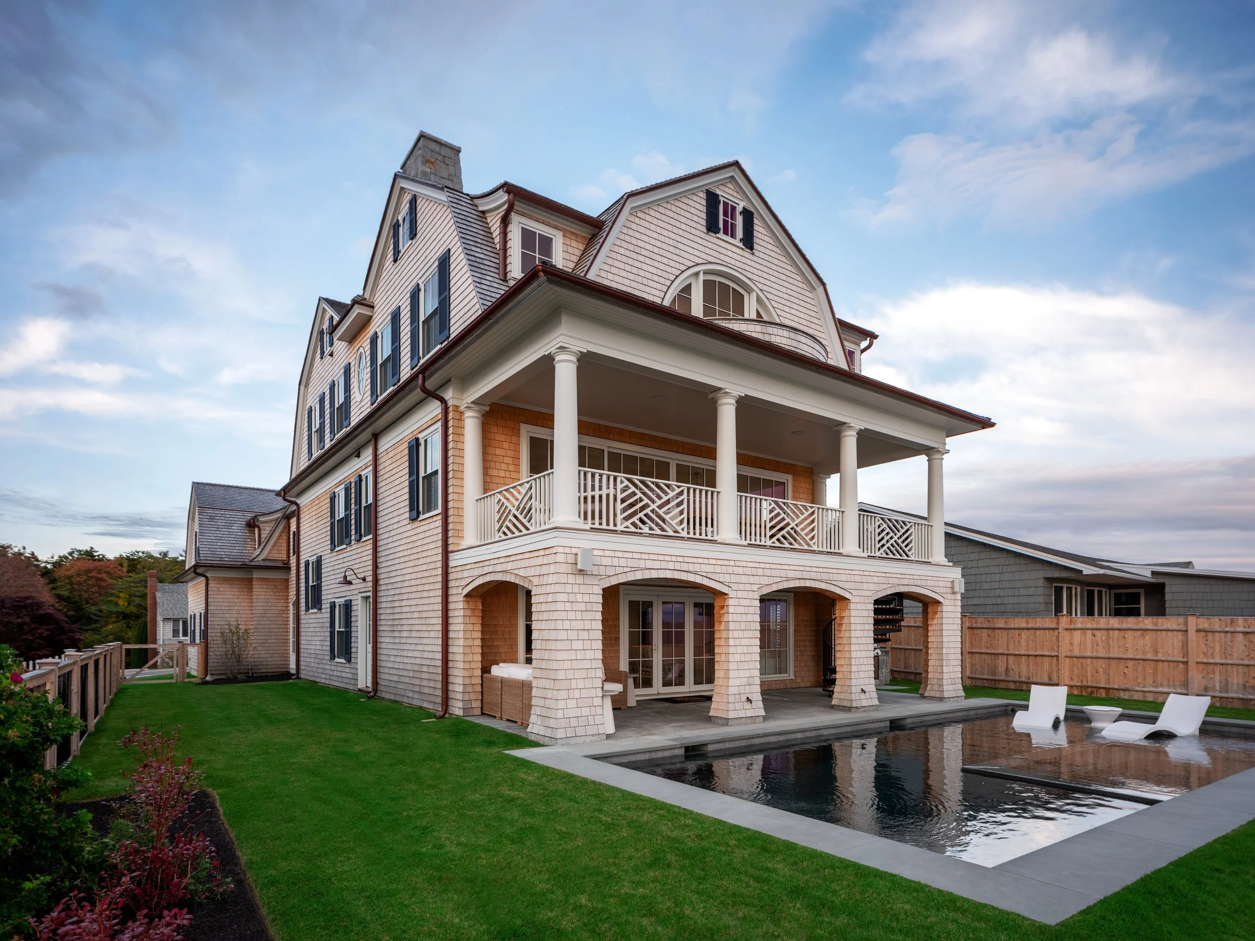 Large multi-story house with a swimming pool in backyard, outdoor furniture, and a wooden fence, during daytime with partly cloudy sky.