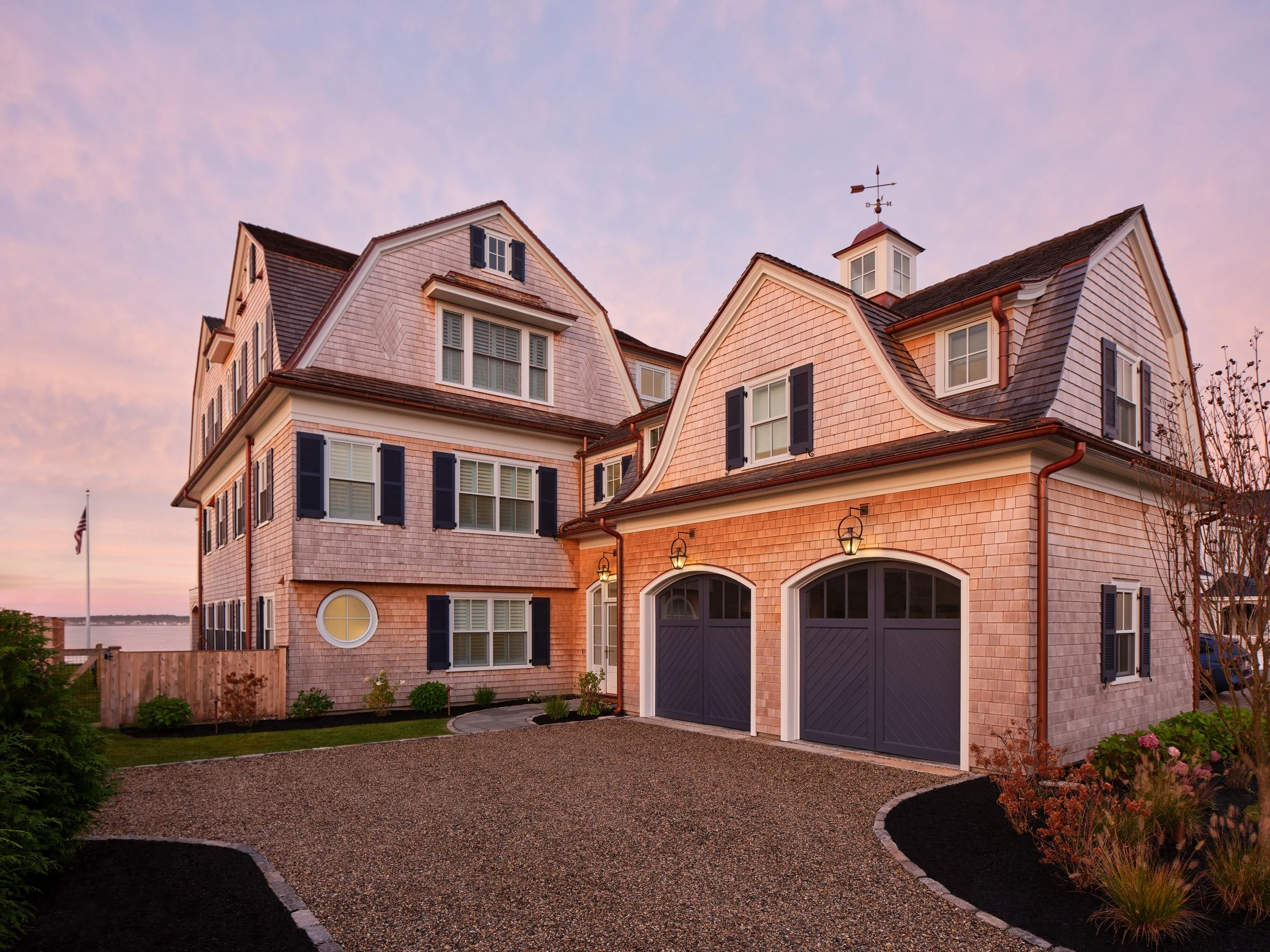 Large traditional house with multiple gables, shingles, and deep blue shutters, at sunset with a pea stone driveway in front and landscaped yard.