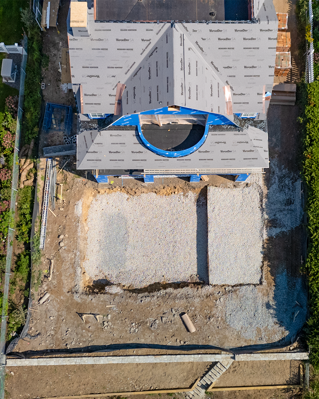 Top-down view of a house under construction, showing the roof with a circular opening, and the surrounding yard with gravel and construction materials.