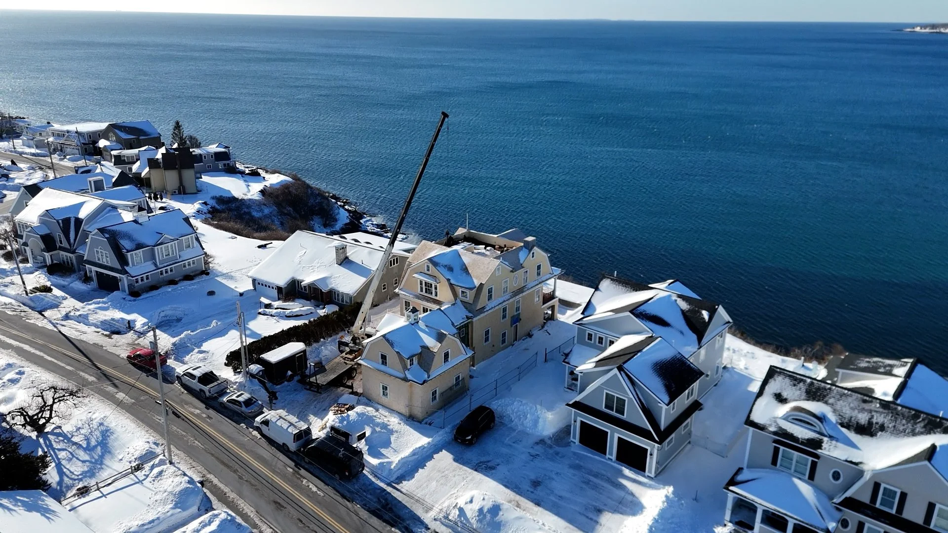 Aerial view of a snowy coastal neighborhood with houses, parked cars, a construction crane, and icy water in the background.
