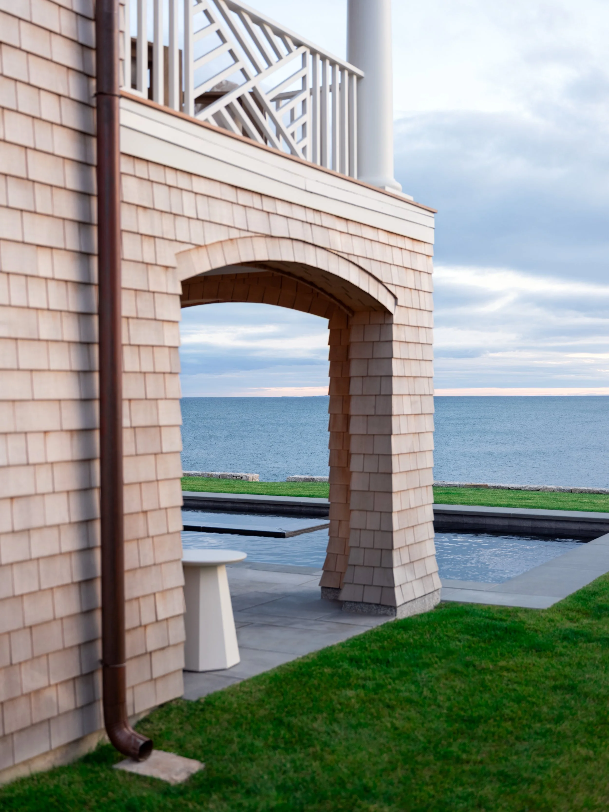 View of a seaside house with an alaskan yellow cedar sided archway and deck, overlooking the ocean, with a grassy yard and a pool in the foreground.