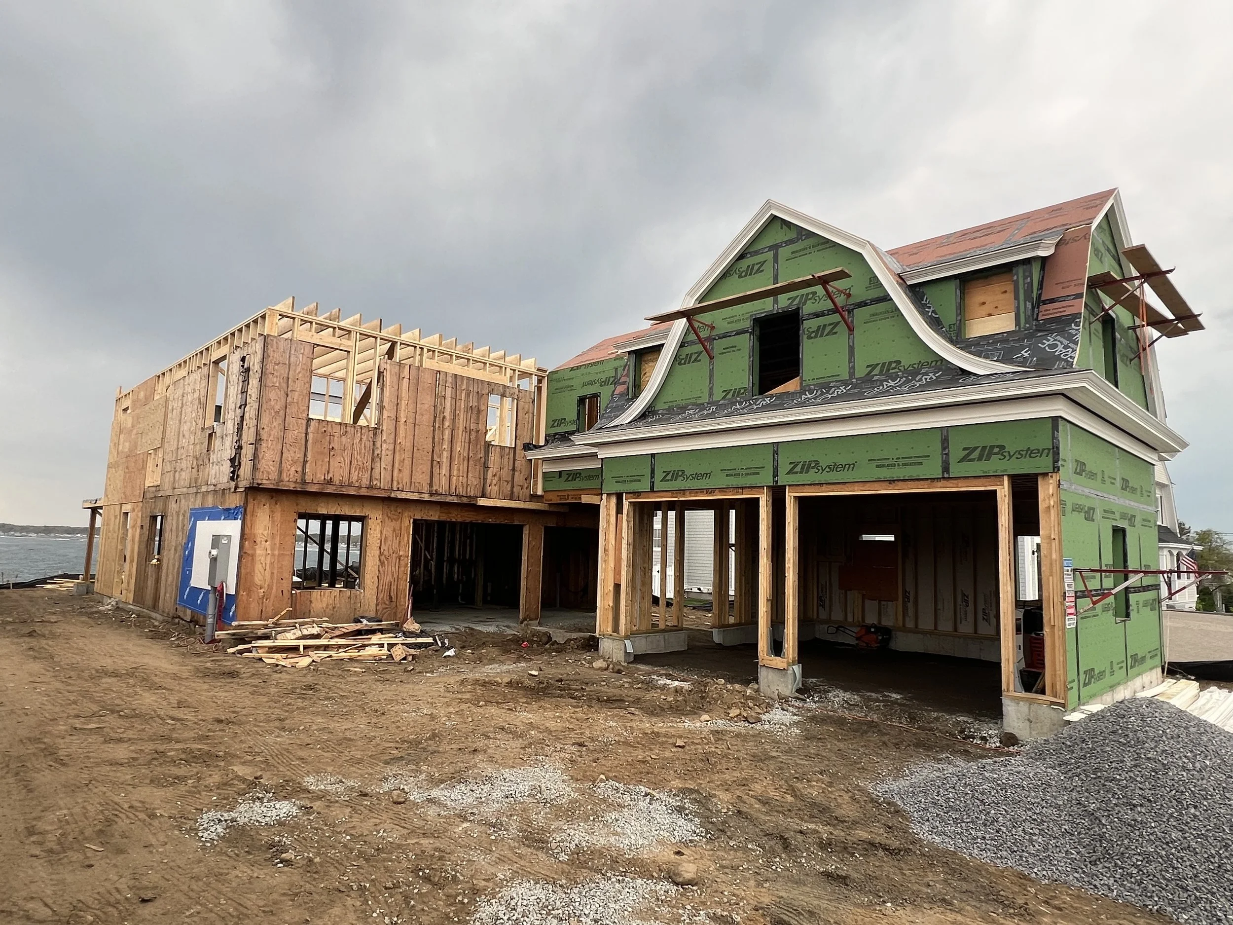 Under construction two-story house with green siding and a mansard roof, situated on a dirt lot, with an adjacent unfinished wooden structure.