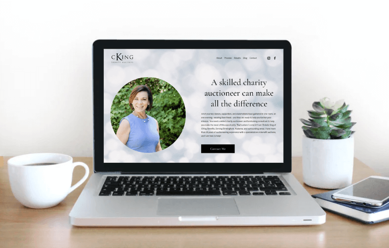 Laptop displaying a website with a portrait of a person and text about charity auctioneering, situated on a desk with a cup of coffee, a smartphone, and a potted plant.