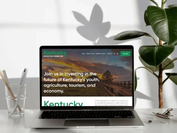 Laptop with Kentucky Exposition Foundation website displayed, surrounded by a pencil holder with pens, a plant, and a pair of glasses.