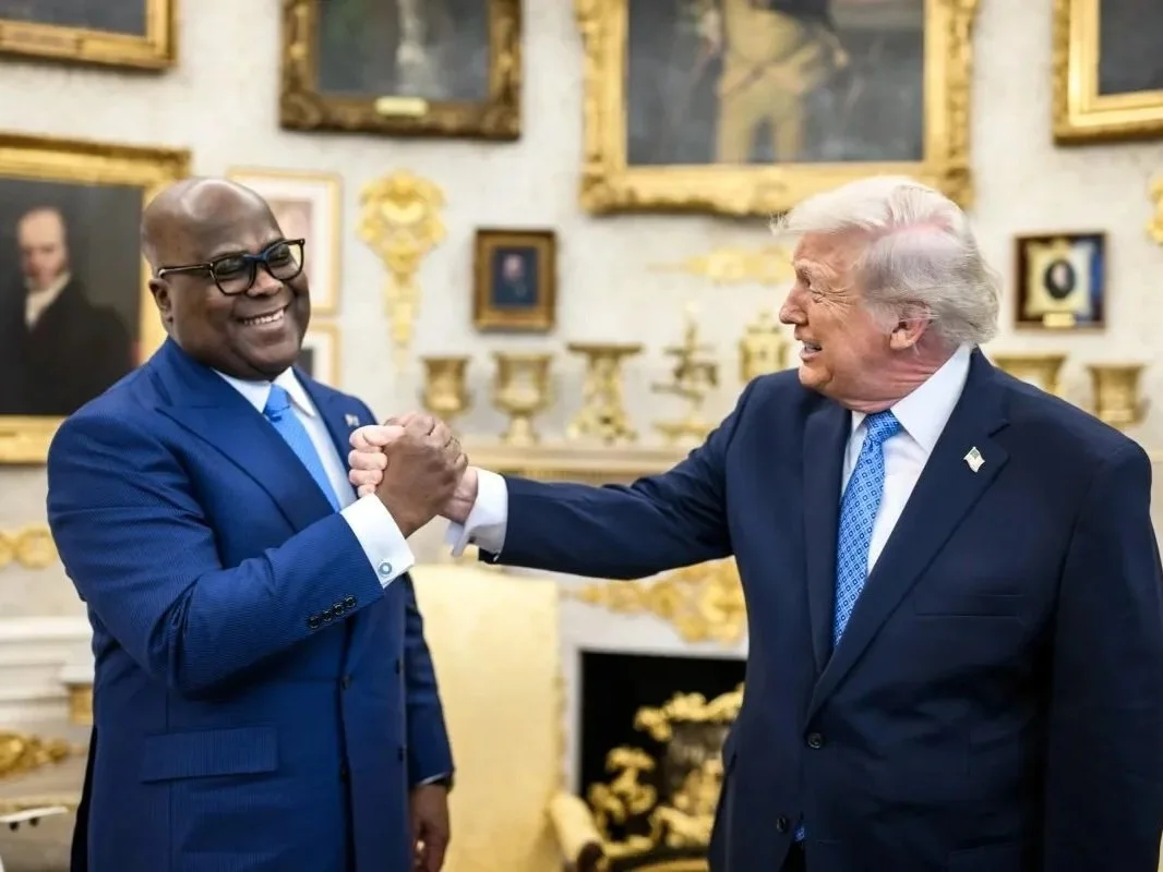 Two men in suits smiling and shaking hands in a gold-accented room with portraits on the wall.