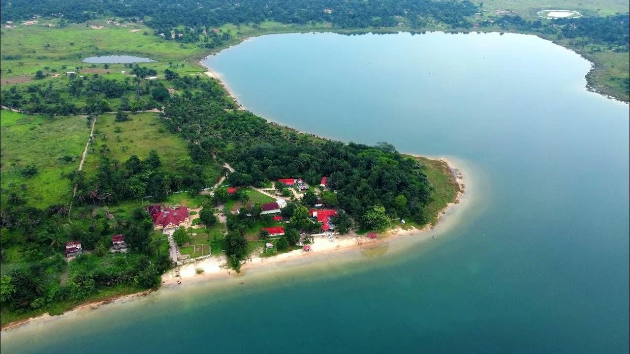 Aerial view of a lakeside area with several buildings, surrounded by greenery and a sandy beach on the shoreline.