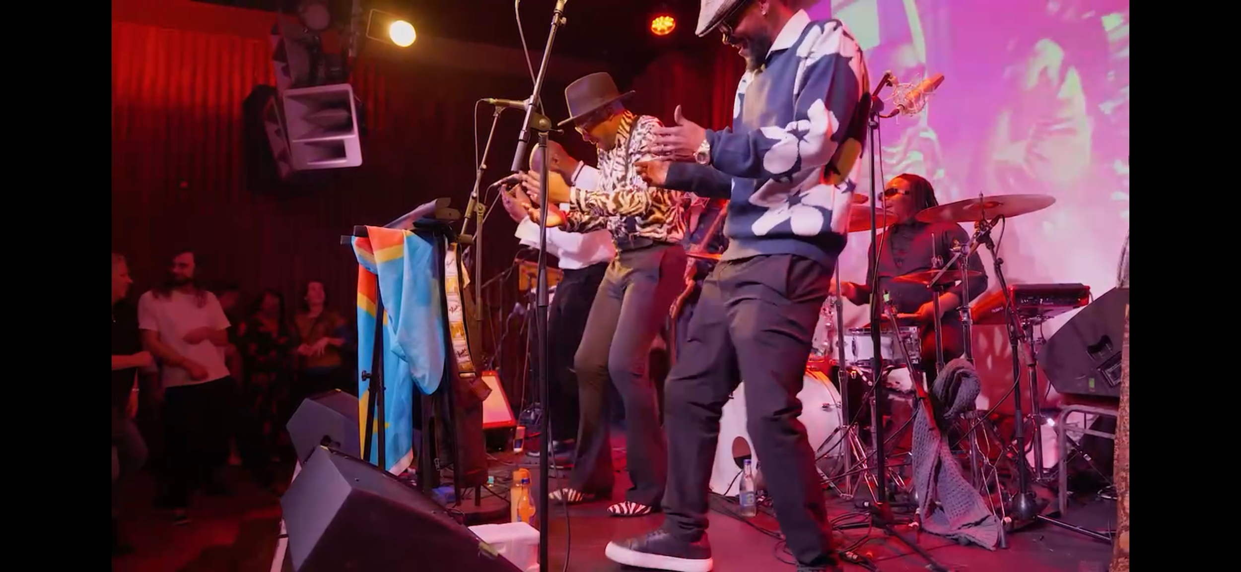Musicians perform on a stage with colorful lighting, including a drummer, a person in a hat, and others, in front of an audience at a live music event.