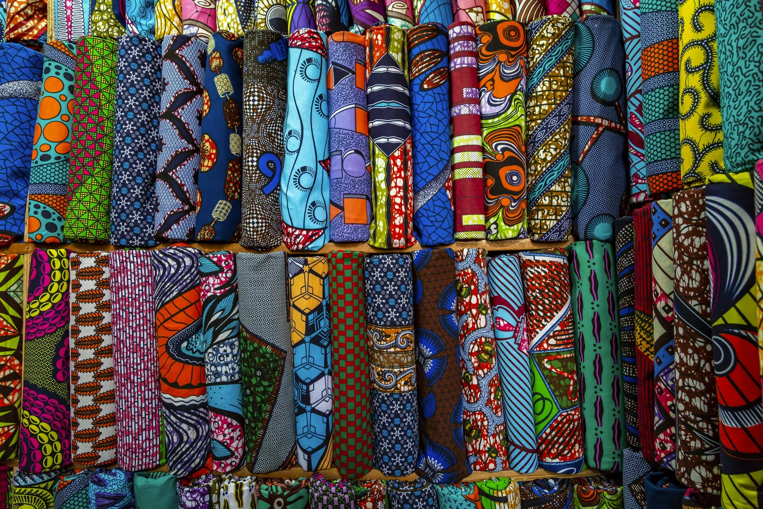 Colorful rolls of African wax print fabric with various intricate patterns displayed on a shelf.