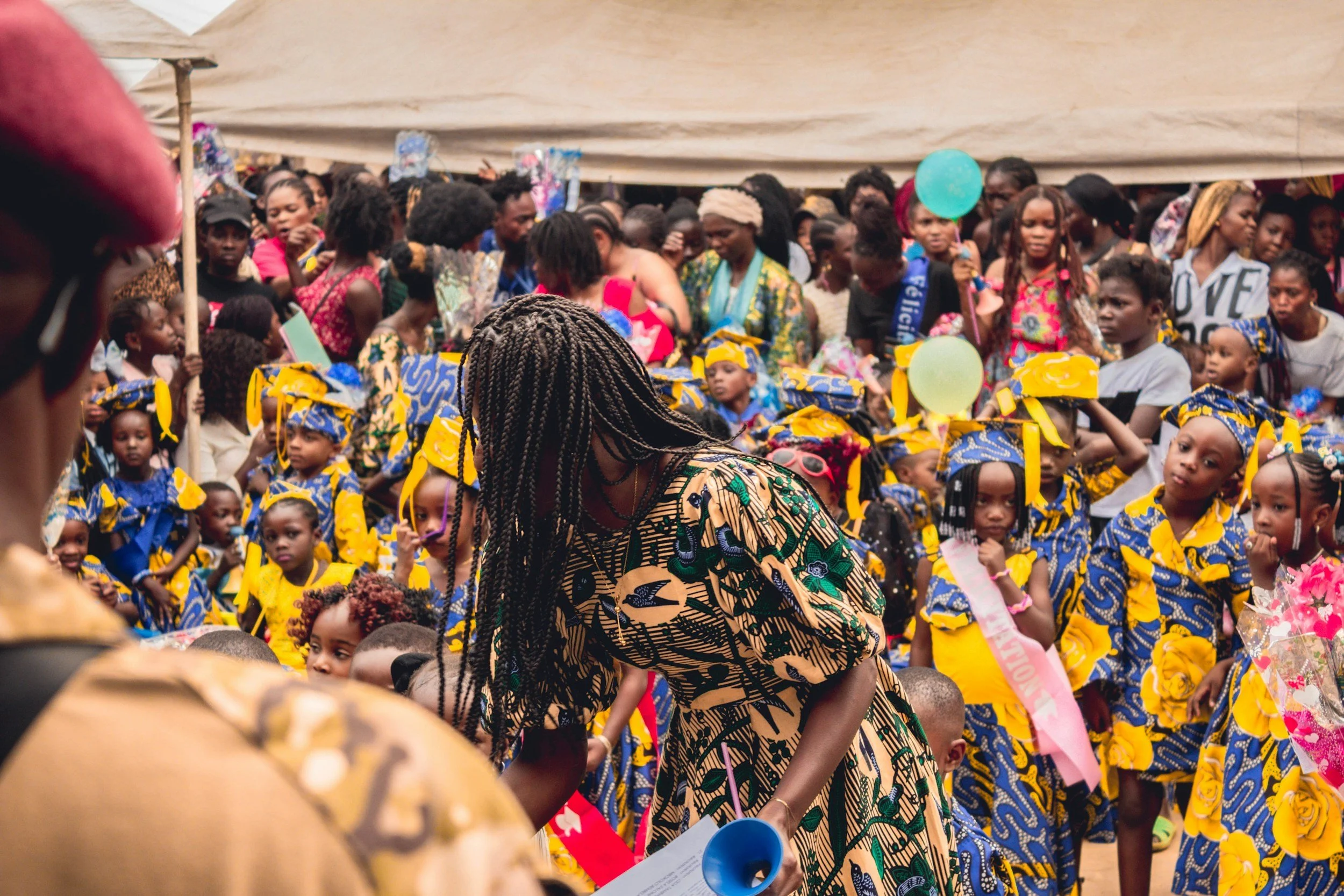 Crowd of children and adults at an outdoor event, some children dressed in matching yellow and blue outfits, with balloons and banners.