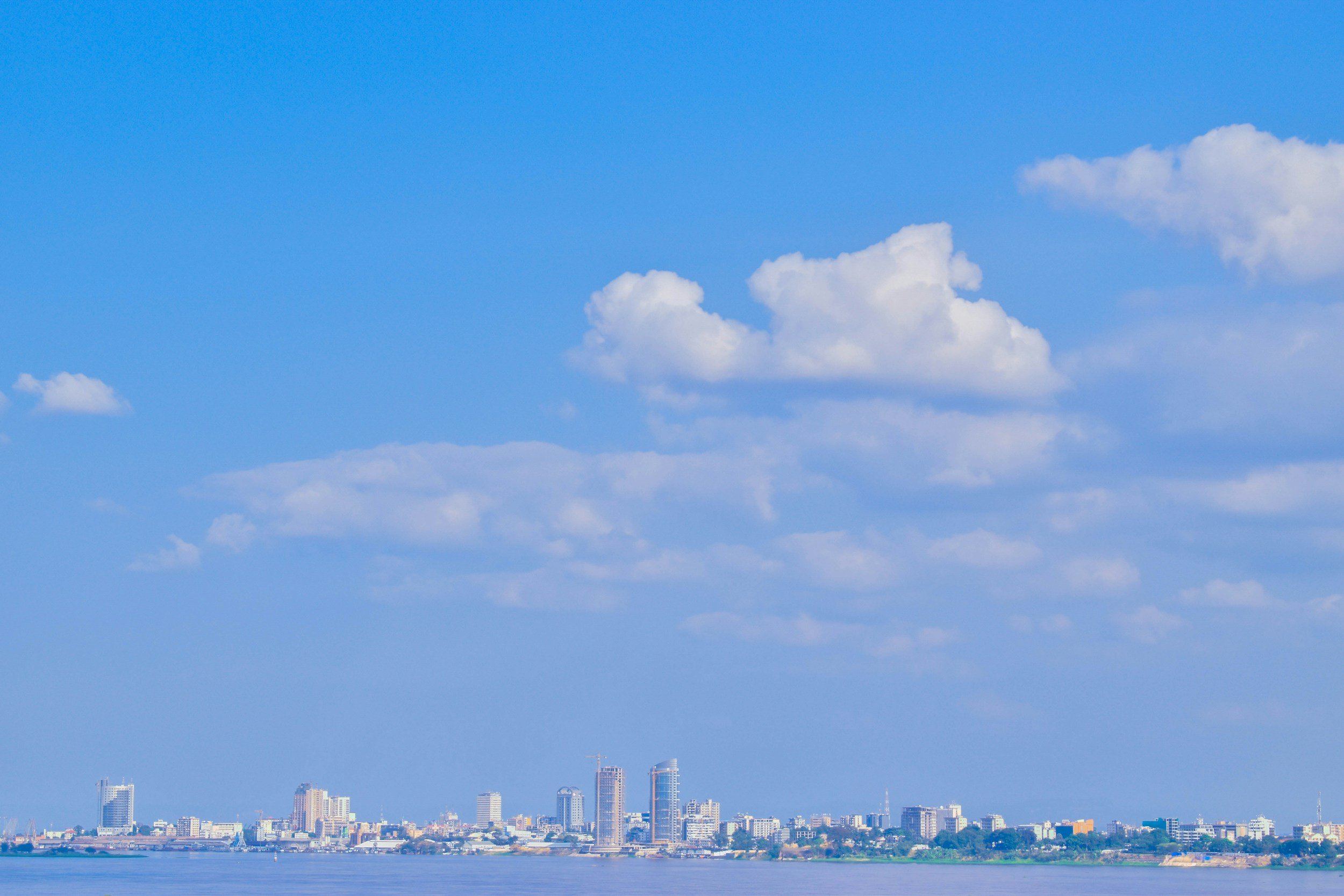 City skyline with a body of water in the foreground and a blue sky with white clouds above.