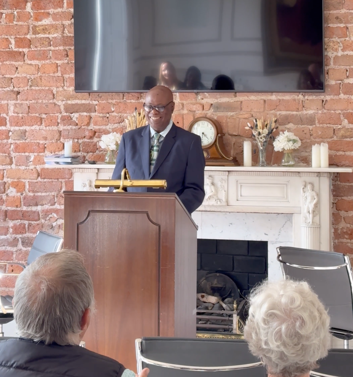 Man in a suit speaking at a podium during a formal event in front of a brick wall with a fireplace and decorative items, while an audience listens.