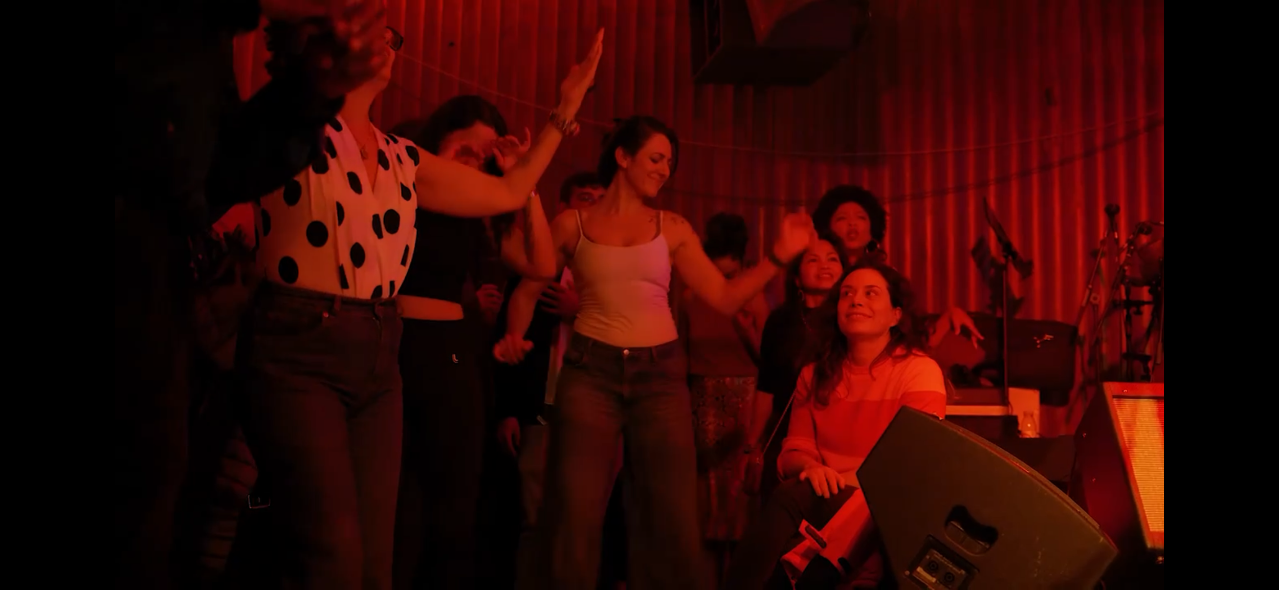 Group of women dancing and singing on stage with red lighting, musical instruments in the background.