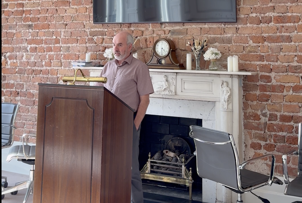 A man standing at a wooden podium in a room with an exposed brick wall, a white fireplace mantle with decorative carvings, a vintage clock, candles, and floral arrangements on the mantle, and a large flat-screen TV mounted above.