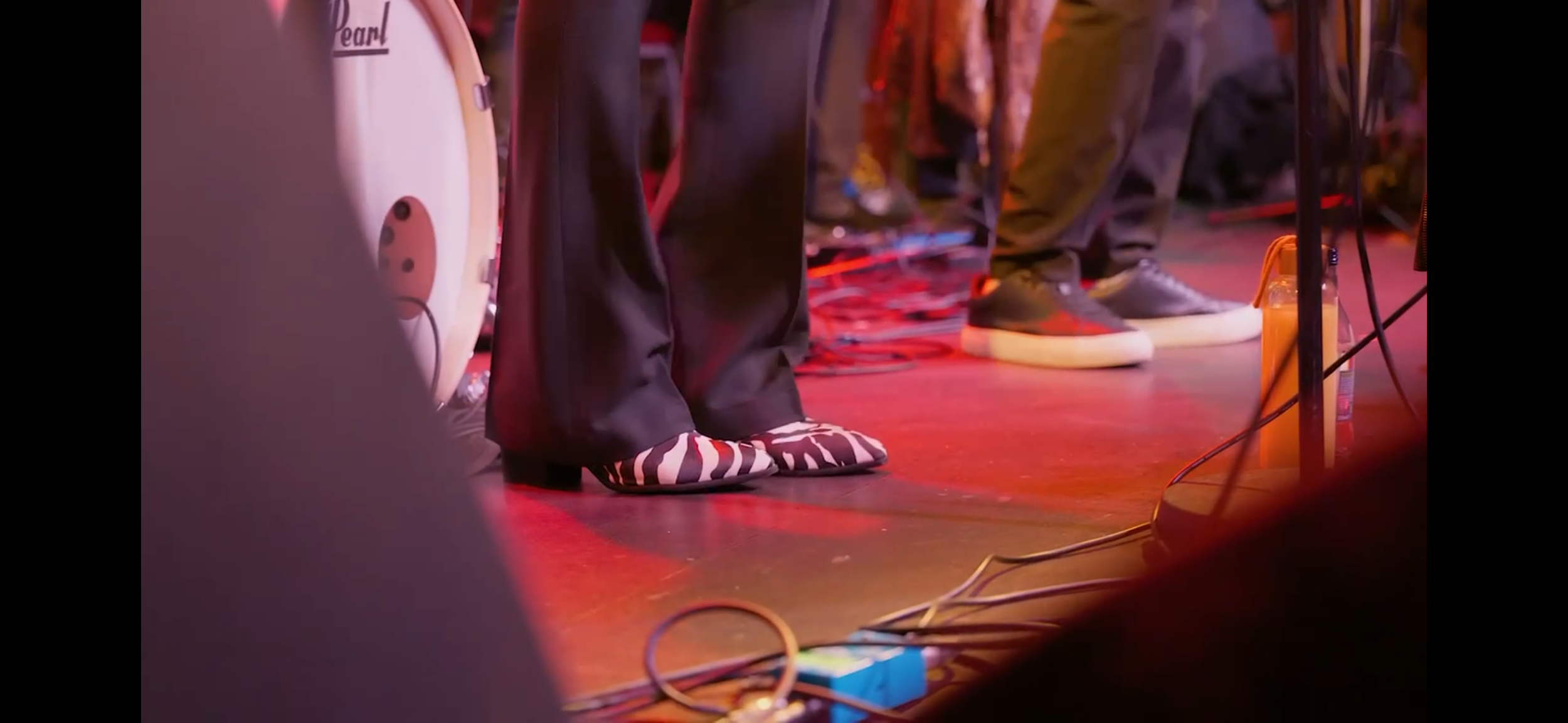 Close-up of feet and lower legs of musicians on stage, with one person wearing zebra-print shoes and others in casual sneakers, surrounded by musical equipment and cables.