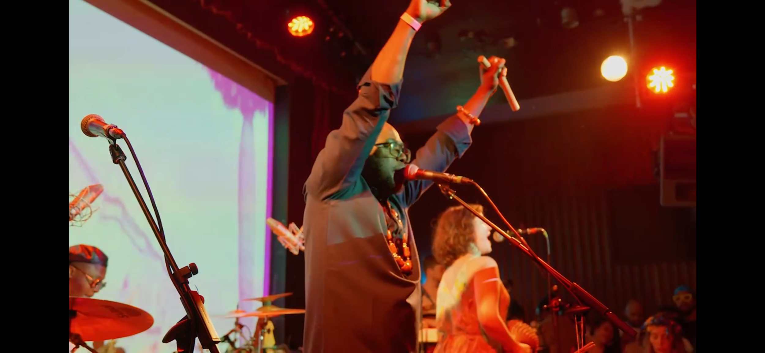 A person with glasses and a black dress is on stage with arms raised, holding a microphone in one hand. They are singing or speaking, with a background of colorful lights and a projection screen. Other band members and audience are visible.