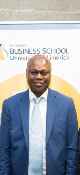 Man in a blue suit and tie standing in front of a Kemmy Business School banner.