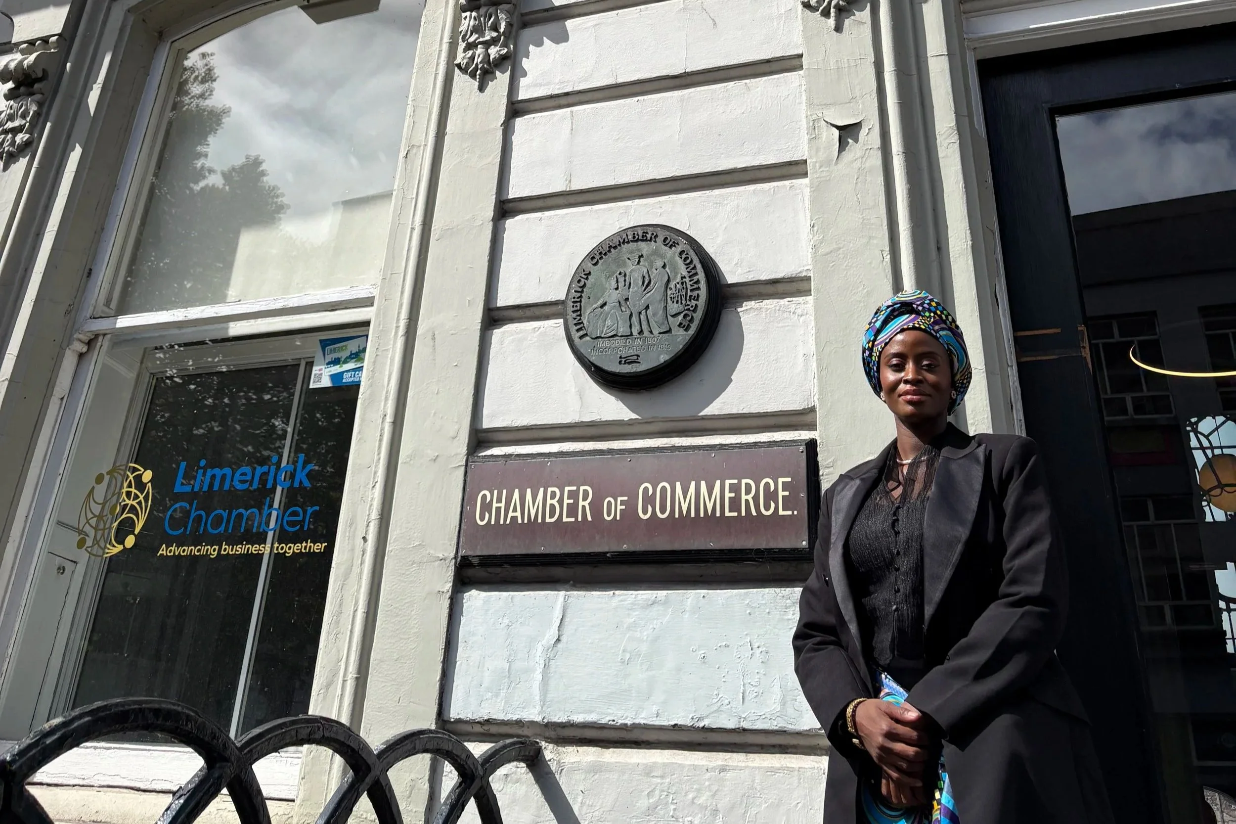 A woman wearing a colorful headwrap and black blazer stands in front of the Chamber of Commerce building in Limerick. The building's exterior has a sign reading 'CHAMBER OF COMMERCE' and a plaque with the city seal. The window to the left features th