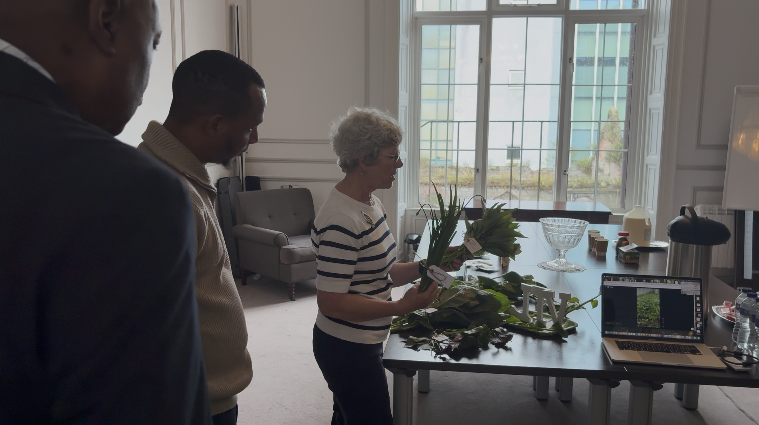 A woman in a striped shirt holding plants and flowers while showing them to two men in a room with large windows, a table with a laptop, glass bowl, and various items on it.