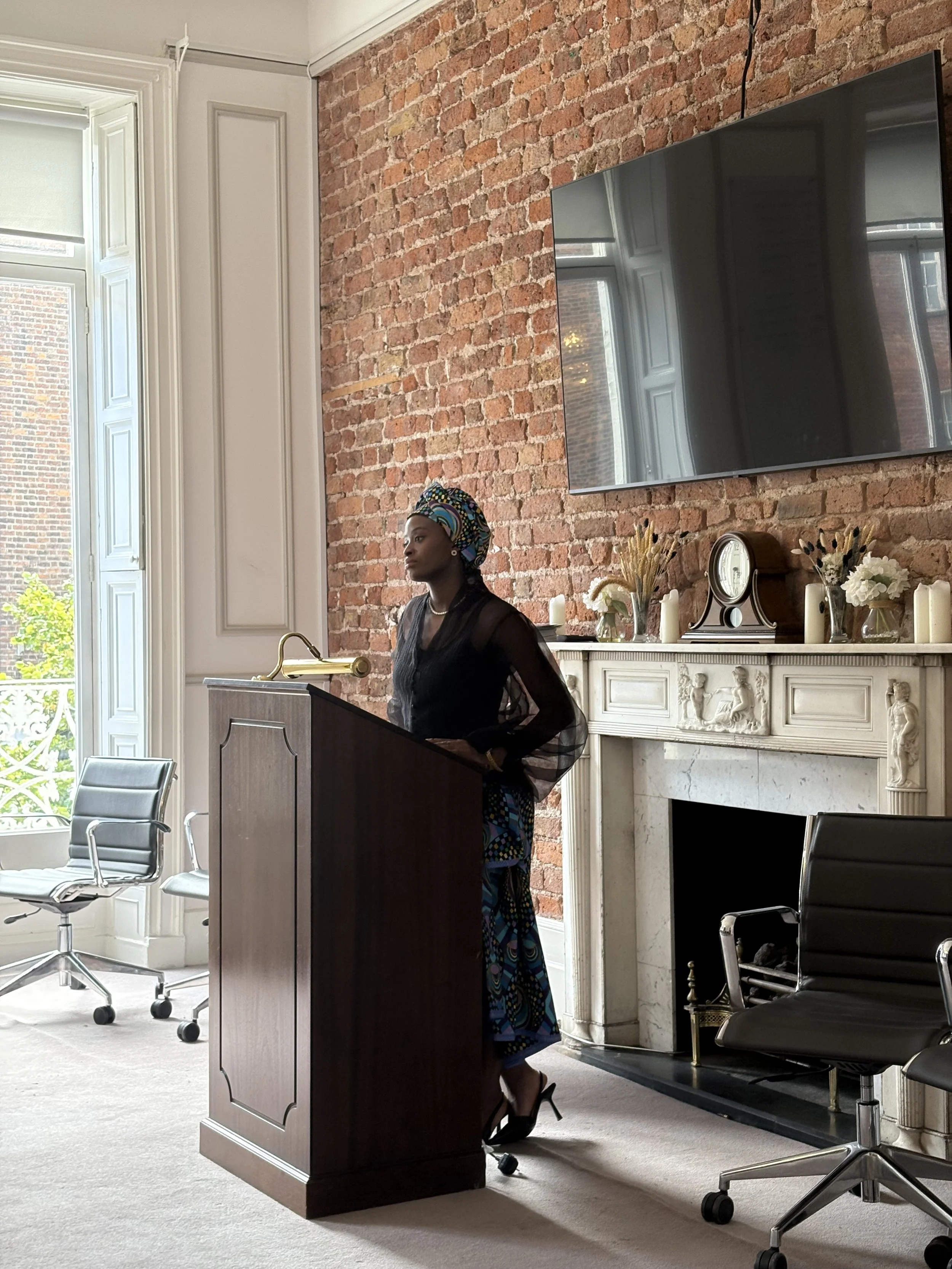 A woman in a colorful headwrap and black sheer top standing at a wooden lectern in a room with exposed brick wall, large window, and ornate fireplace. There are black office chairs and a large flat-screen TV on the wall.