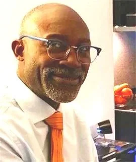 A middle-aged man with glasses and a beard, wearing a white shirt and an orange tie, smiling in an indoor setting.