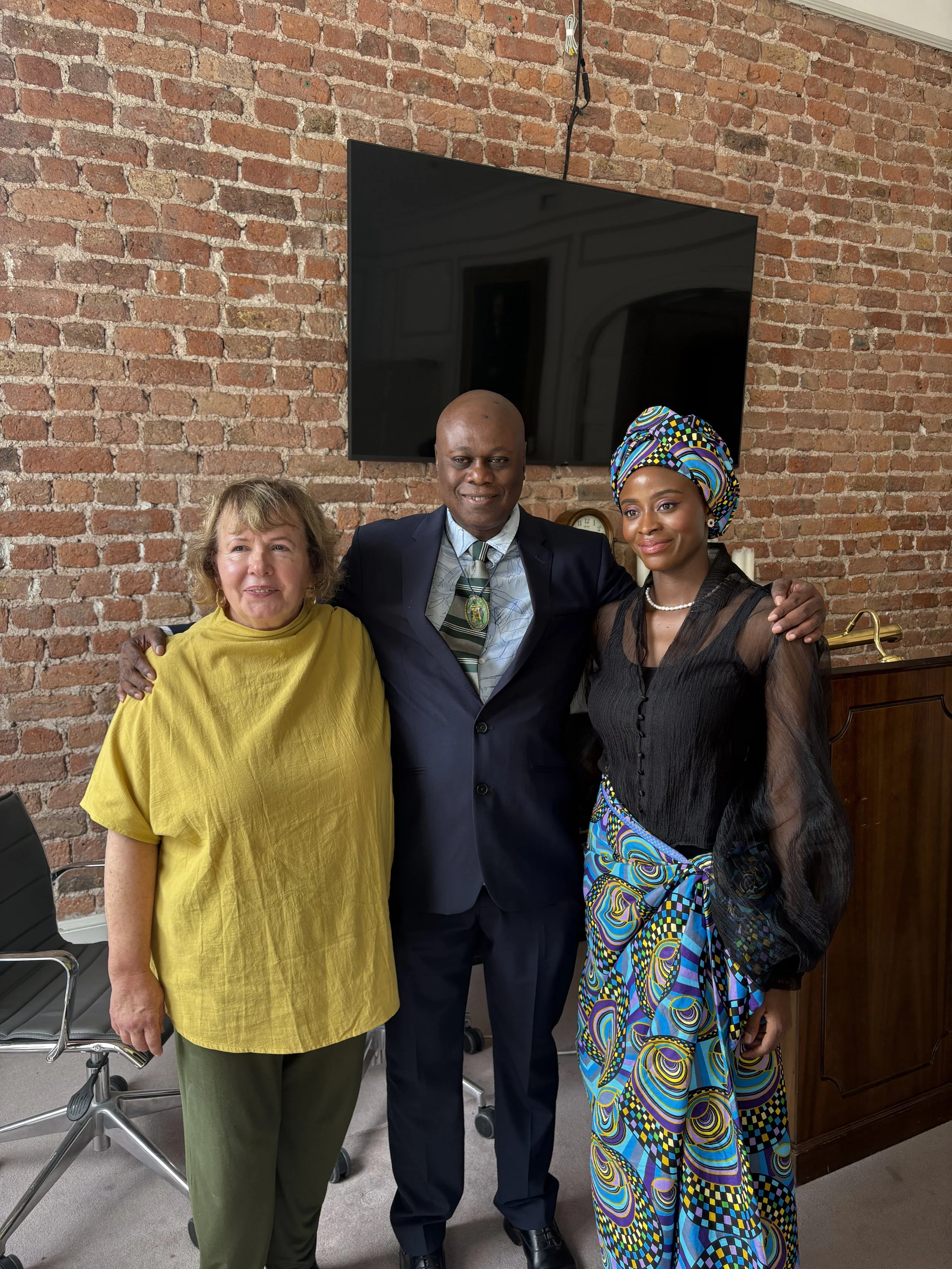 Three people standing together indoors in front of a brick wall and a large black television screen, smiling.