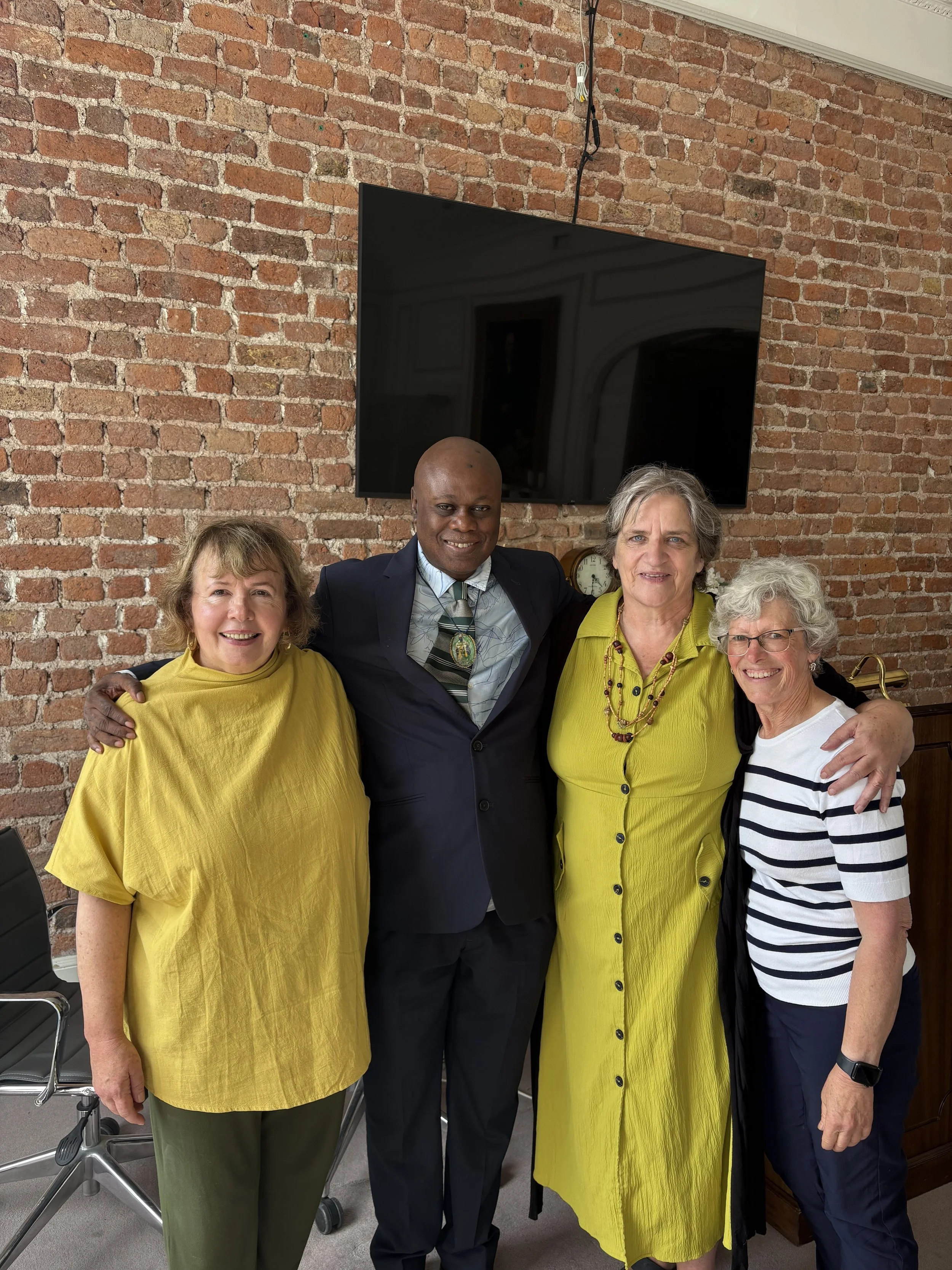 Four people standing together, smiling, with a brick wall and a TV screen behind them.