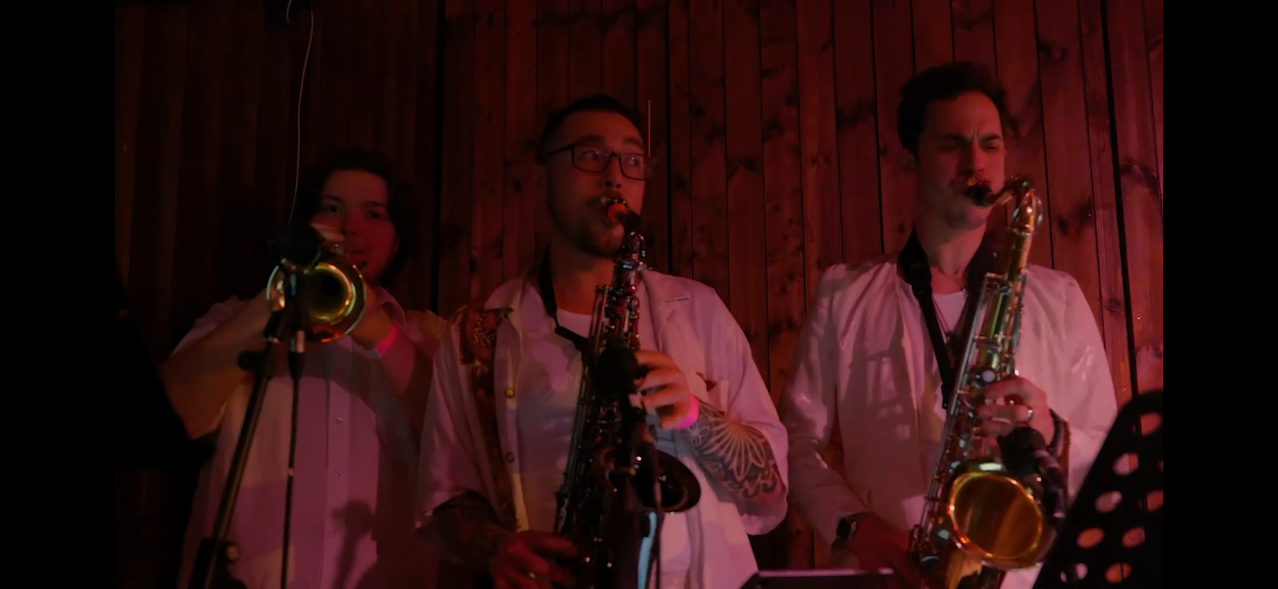 Three musicians playing saxophones in front of a wooden wall, with red lighting.
