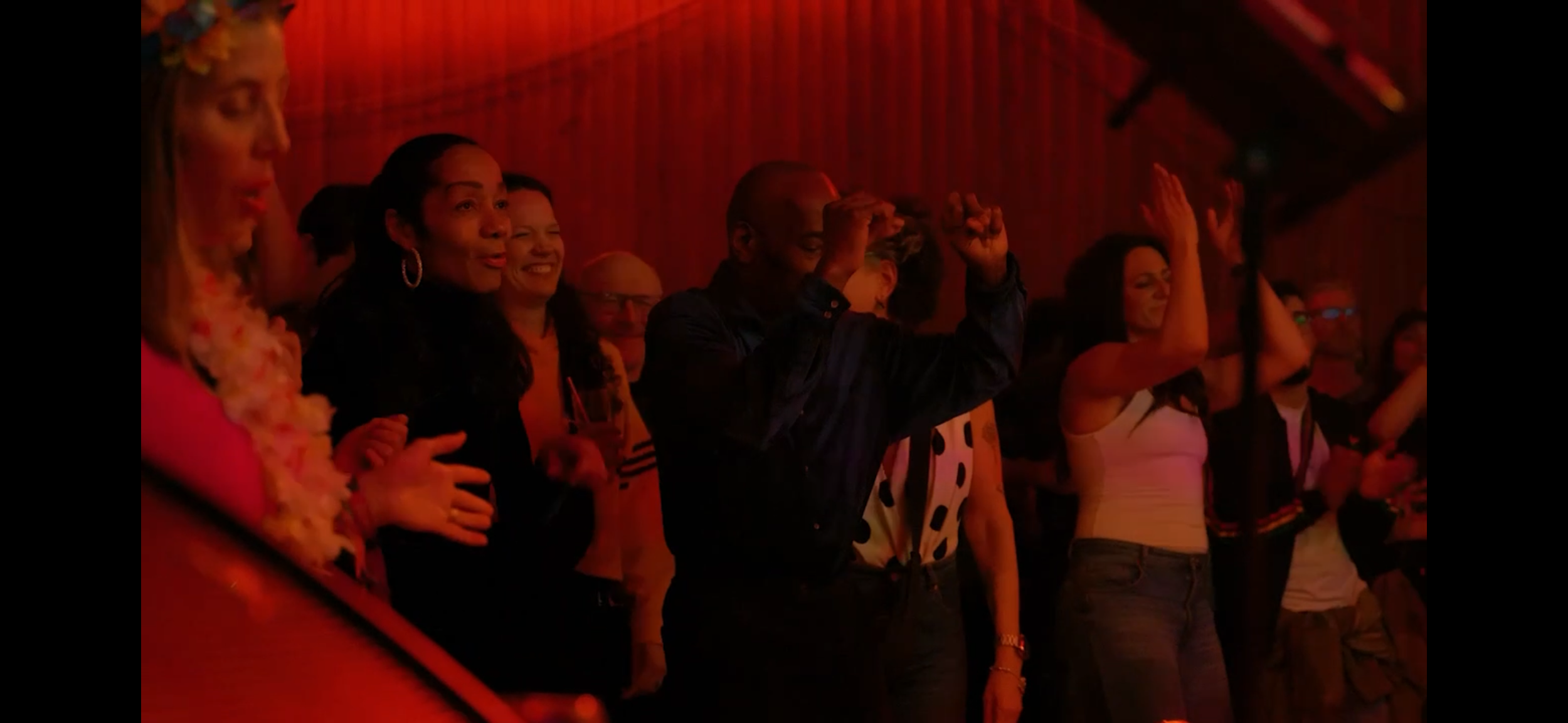 Group of people at an indoor event, some with hands raised, in dim red lighting.
