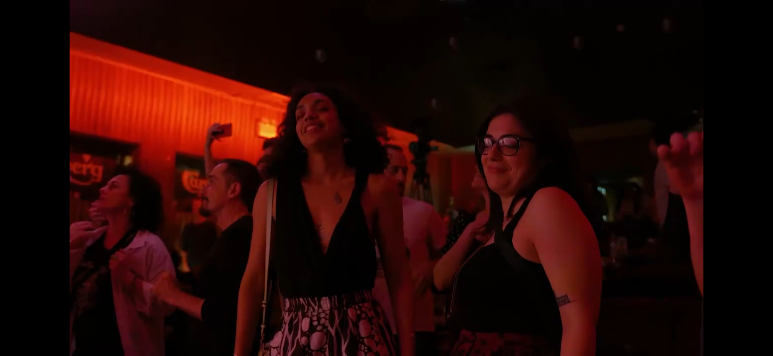 Group of friends dancing and smiling at a bar or nightclub, with red lighting and music posters on the wall.