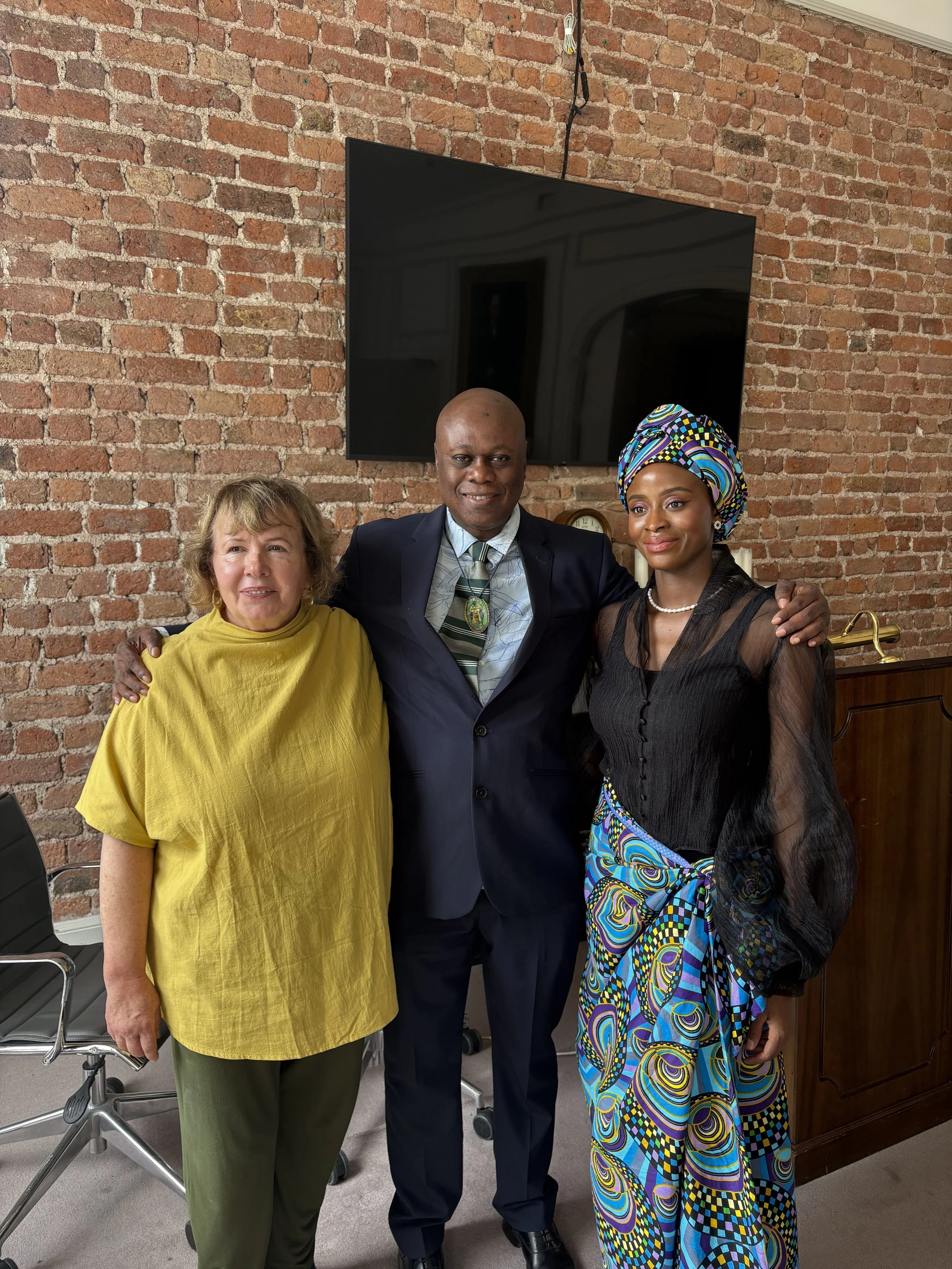 Three people standing in an office with a brick wall, a large television, and a wooden desk. The woman on the left has light skin and short light brown hair, wearing a yellow top and green pants. The man in the middle has dark skin and is bald, wearing a dark suit, white shirt, striped tie, and a Kenyan badge on his suit. The woman on the right has dark skin, wearing a colorful patterned headwrap, black sheer blouse, and matching patterned skirt, with a pearl necklace.