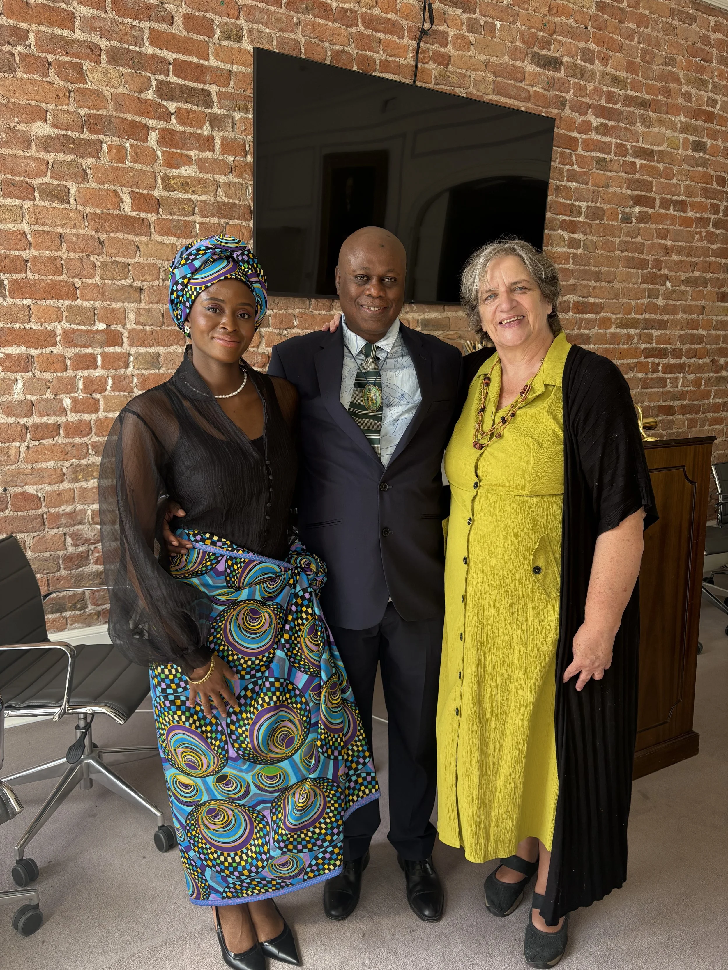 Three people standing together in an office with a brick wall and a large flat-screen TV behind them. The woman on the left wears African traditional clothing and headwrap with a colorful pattern. The man in the middle wears a dark suit and tie with 