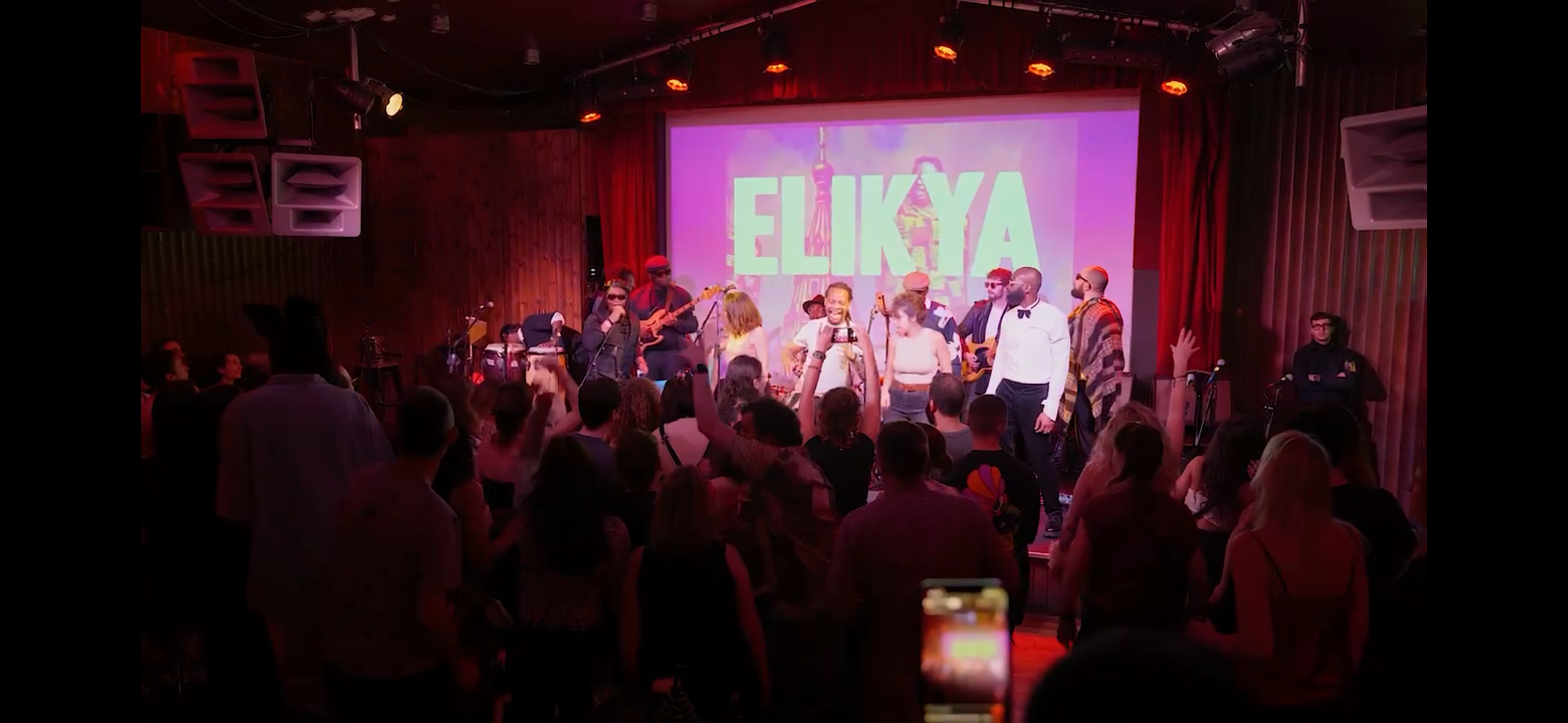 A live performance on stage with musicians and dancers, and an audience in a dimly lit venue with wooden walls and a large screen displaying the word 'ELIKYA' in the background.