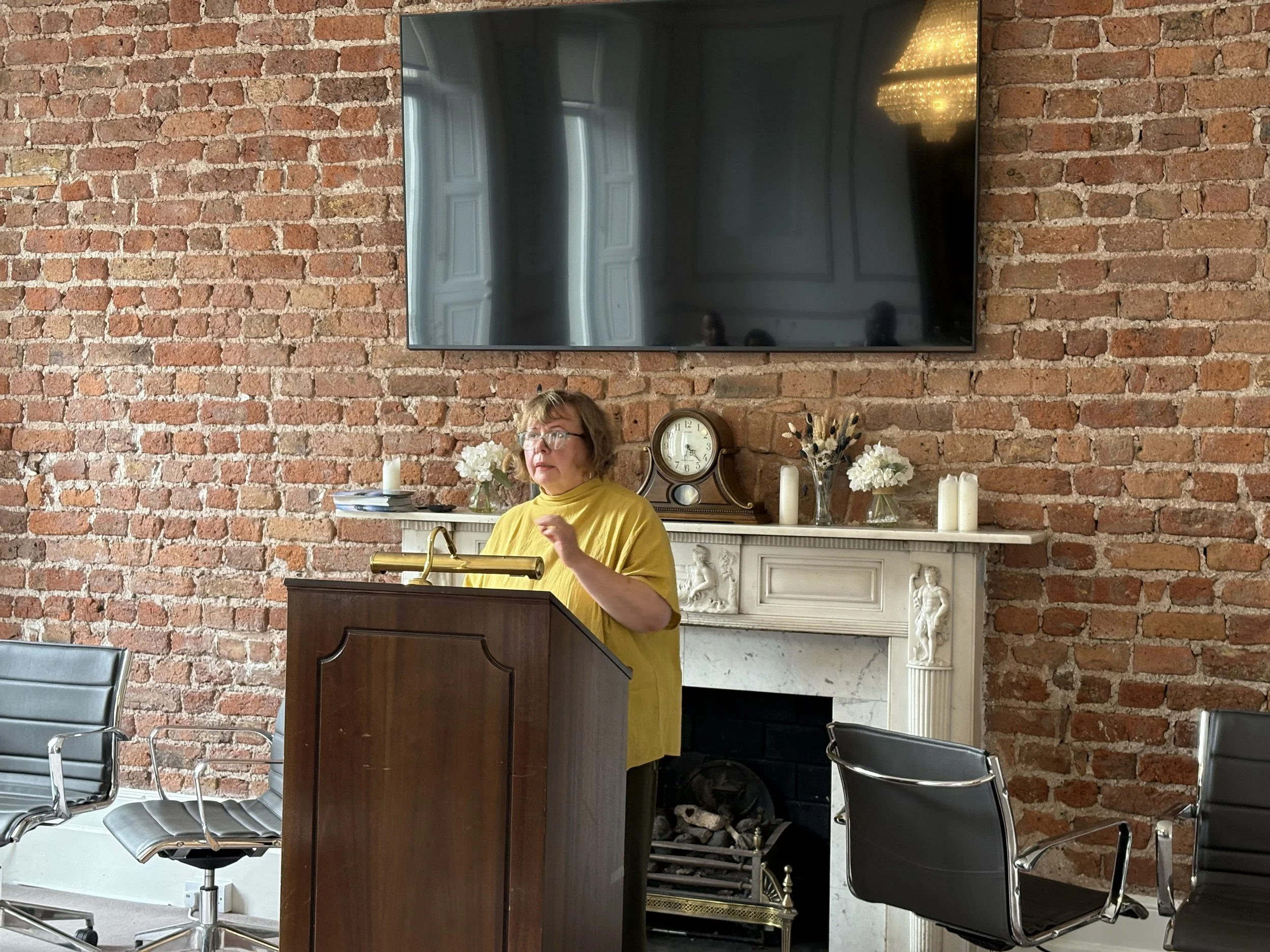 Woman in yellow shirt giving a speech at a wooden podium in a room with an exposed brick wall, a fireplace with decorative photos and candles, and a large TV mounted on the wall.