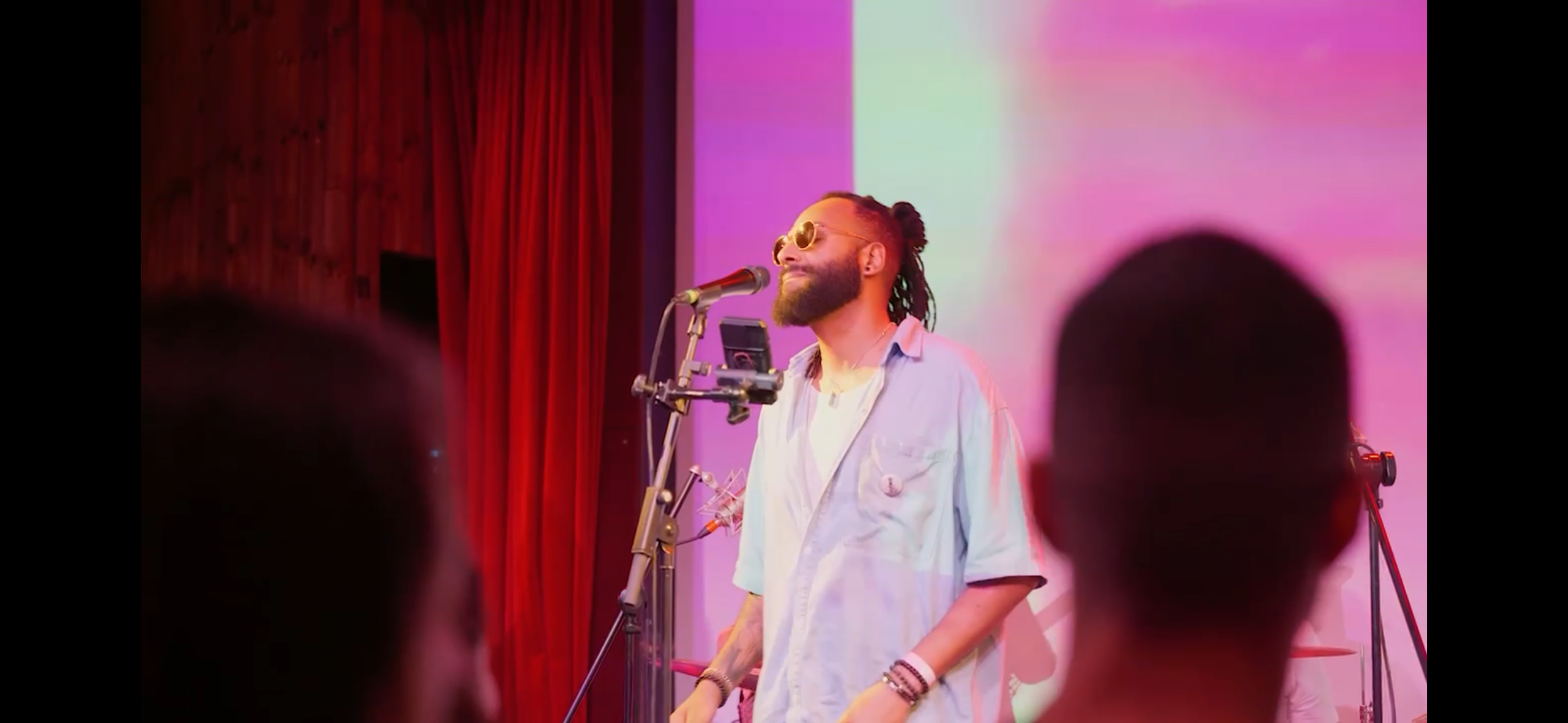 A man with dreadlocks, beard, and sunglasses smiles on stage with a microphone, wearing a light-colored shirt, with a colorful background and red curtains behind him.