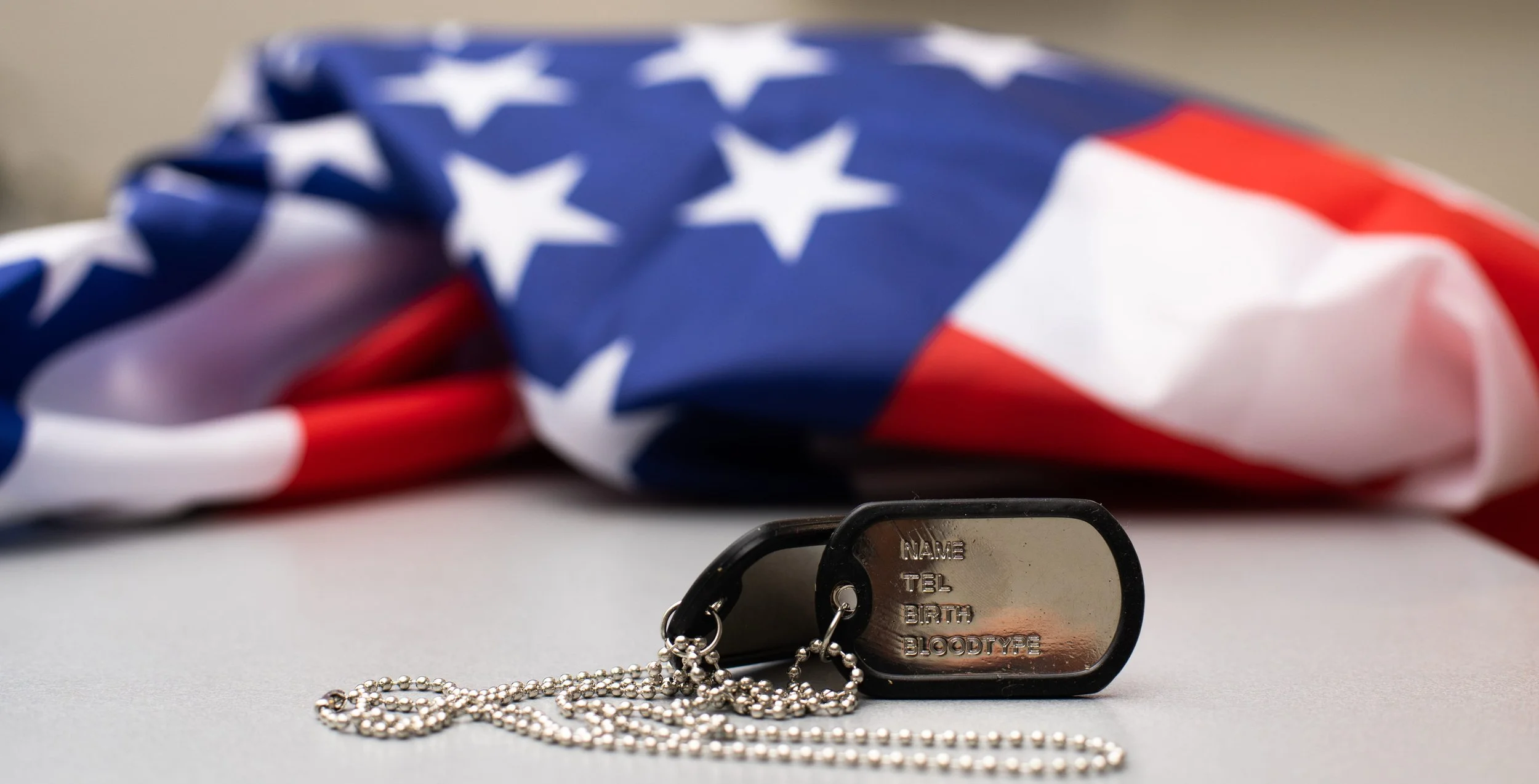 A U.S. flag lying flat with a dog tag in front of it. The dog tag has spaces labeled for personal information.