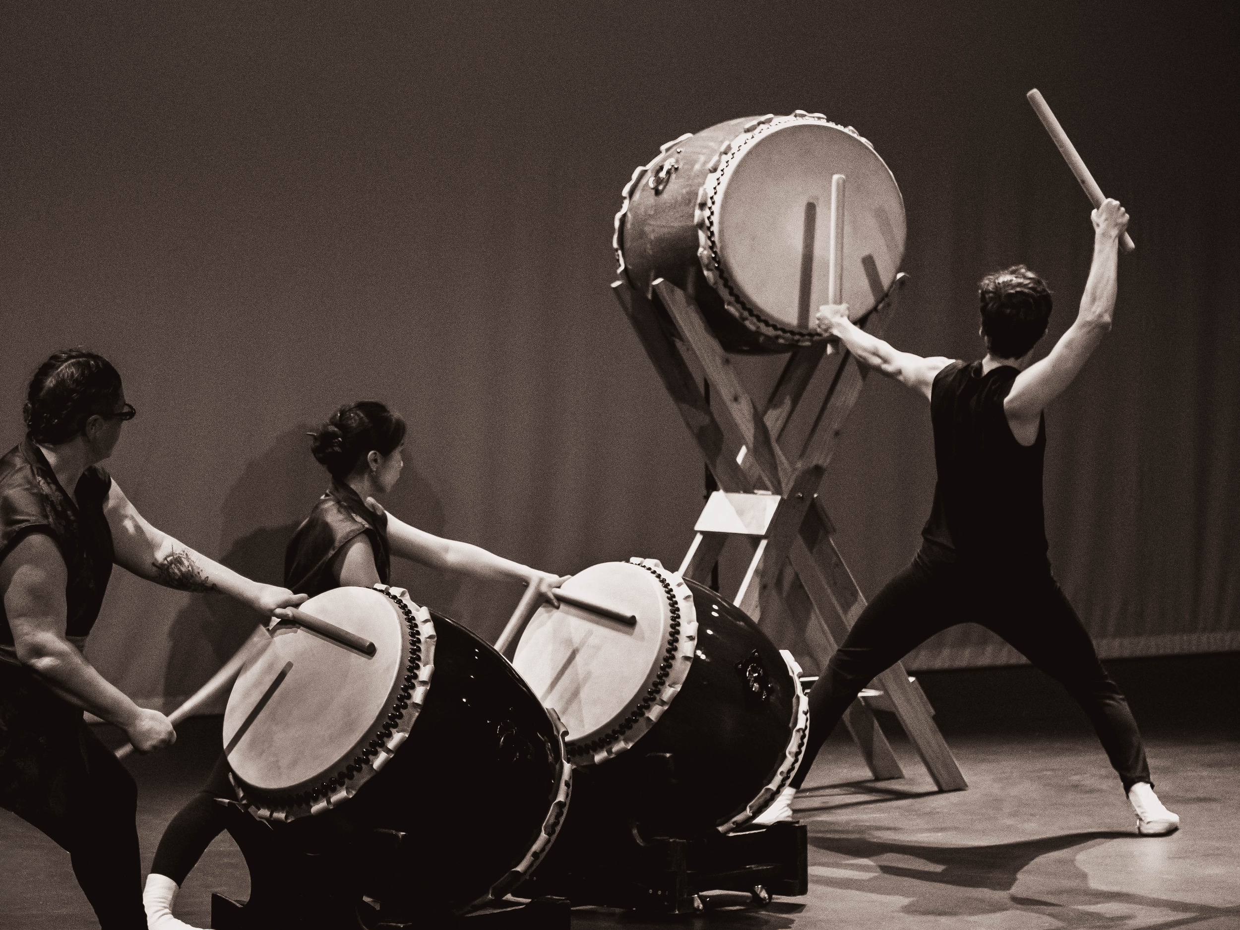 Men and women playing traditional Japanese taiko drums on stage, with one man standing and striking a large drum with drumsticks. The scene is in black and white.