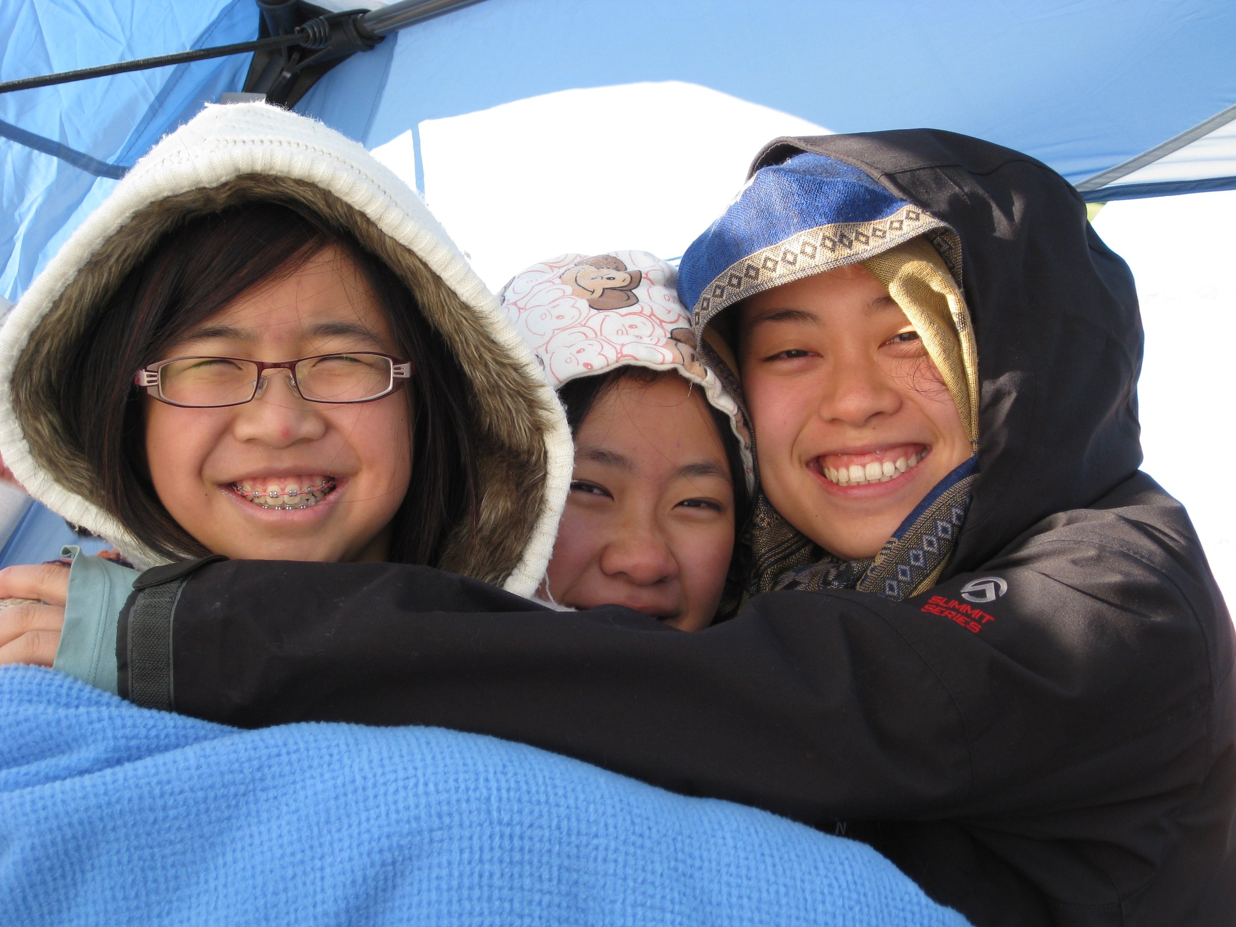 Three smiling girls wearing winter jackets and hats inside a blue tent, hugging each other.