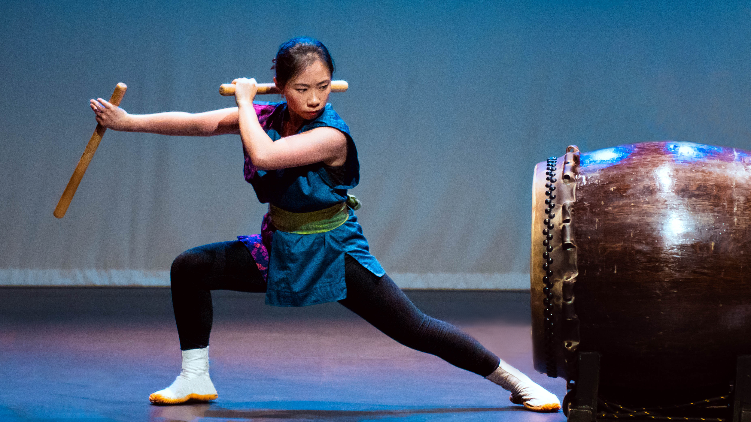 A young woman in traditional Asian attire plays a large drum with a wooden stick on stage.