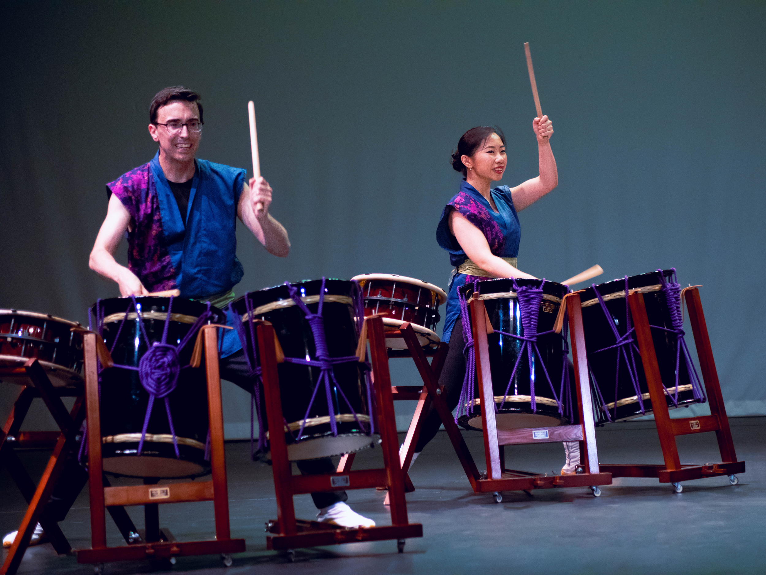 Two people playing traditional Japanese drums on stage. They are wearing blue and purple outfits and appear to be performing energetically.
