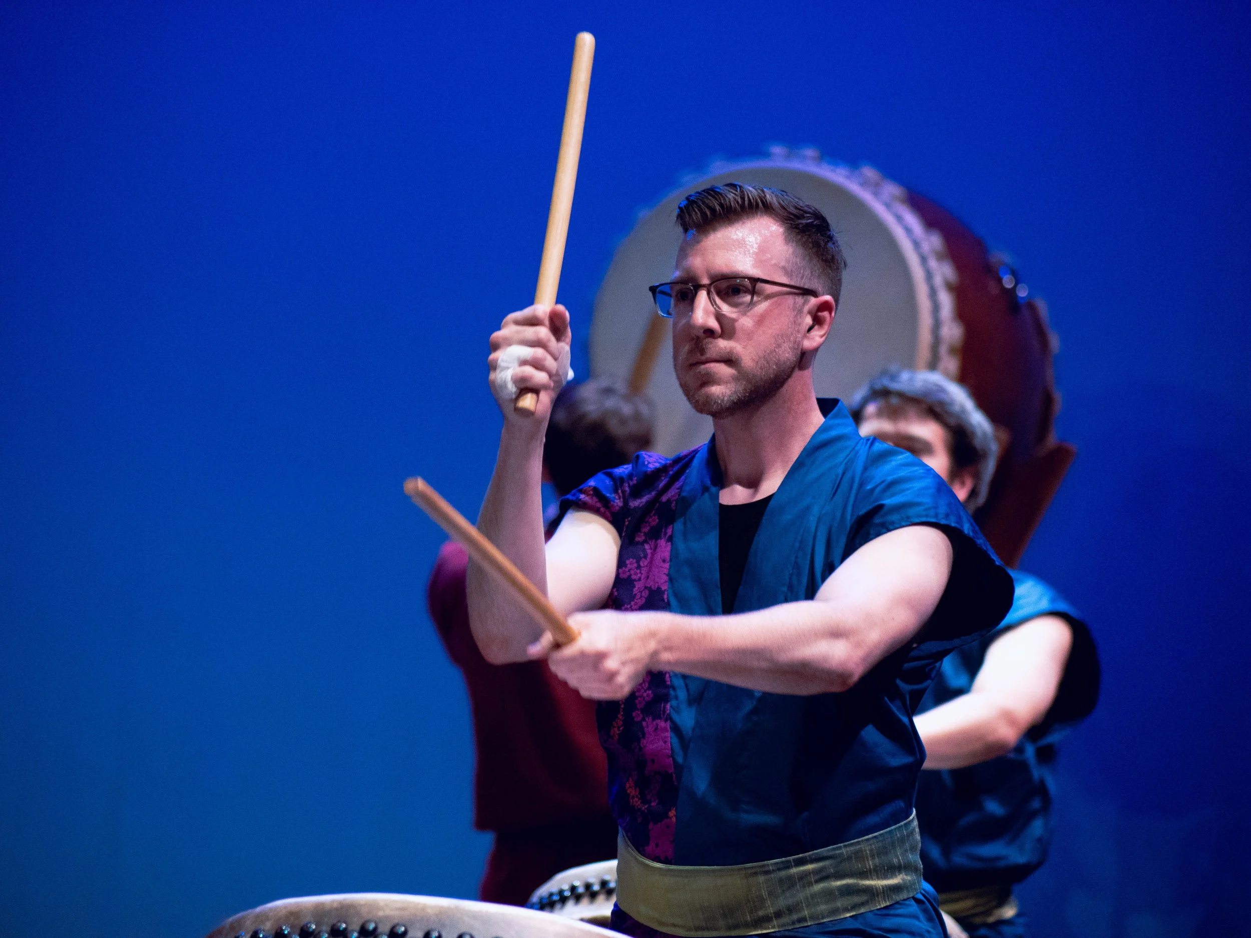 Man playing large traditional drum with drumsticks, wearing glasses and a blue shirt, on stage with other performers in the background