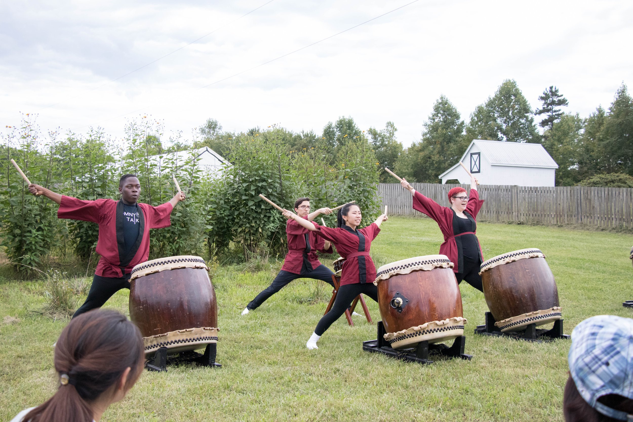Four people playing traditional Japanese taiko drums outdoors on grass, dressed in maroon robes, with a backdrop of trees, a fence, and a white shed. Audience members are visible in the foreground.